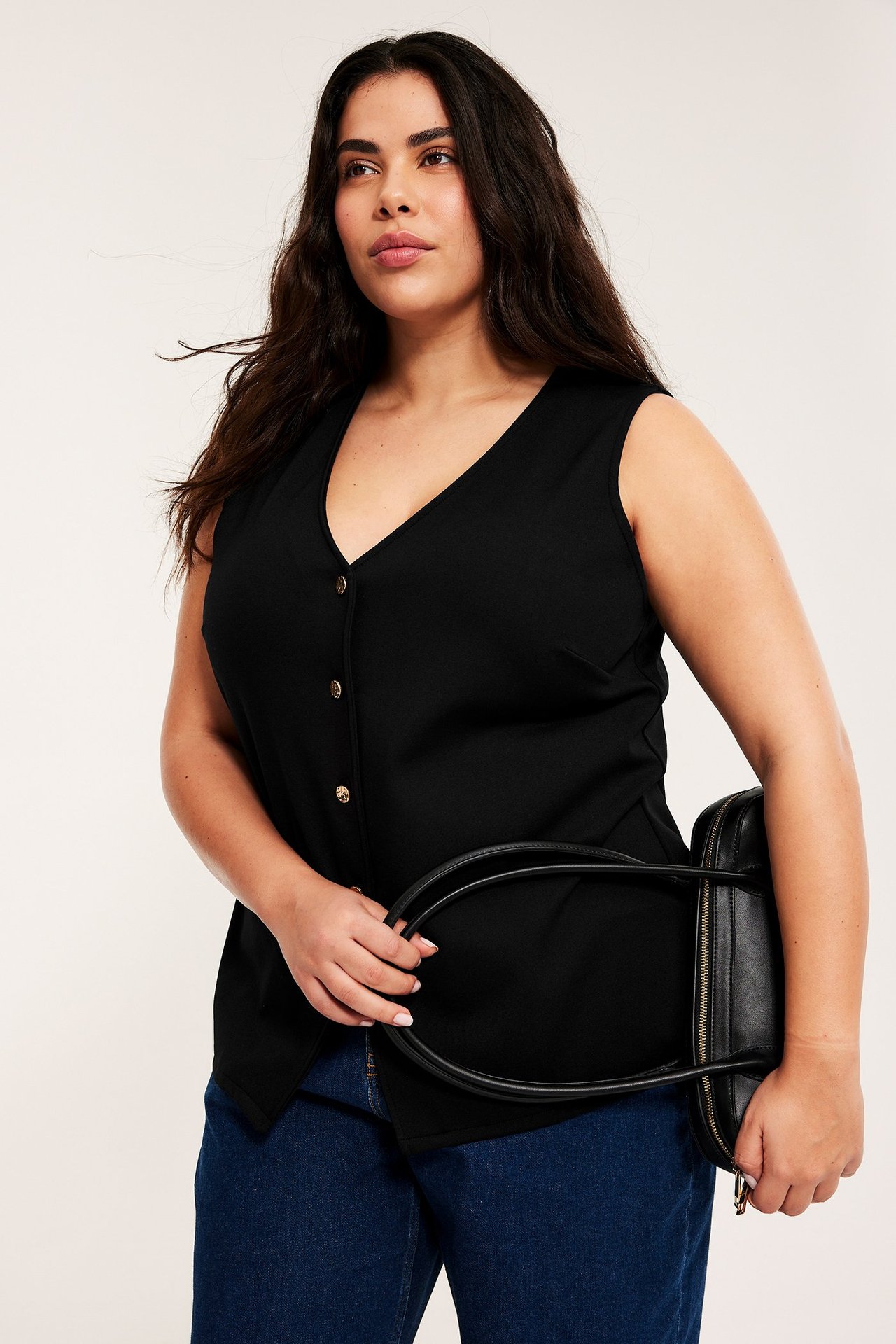 Naisen etunäkymä mustassa trikoovestissä kultaisilla napeilla, sinisissä farkuissa ja mustaa käsilaukkua pitelemässä.