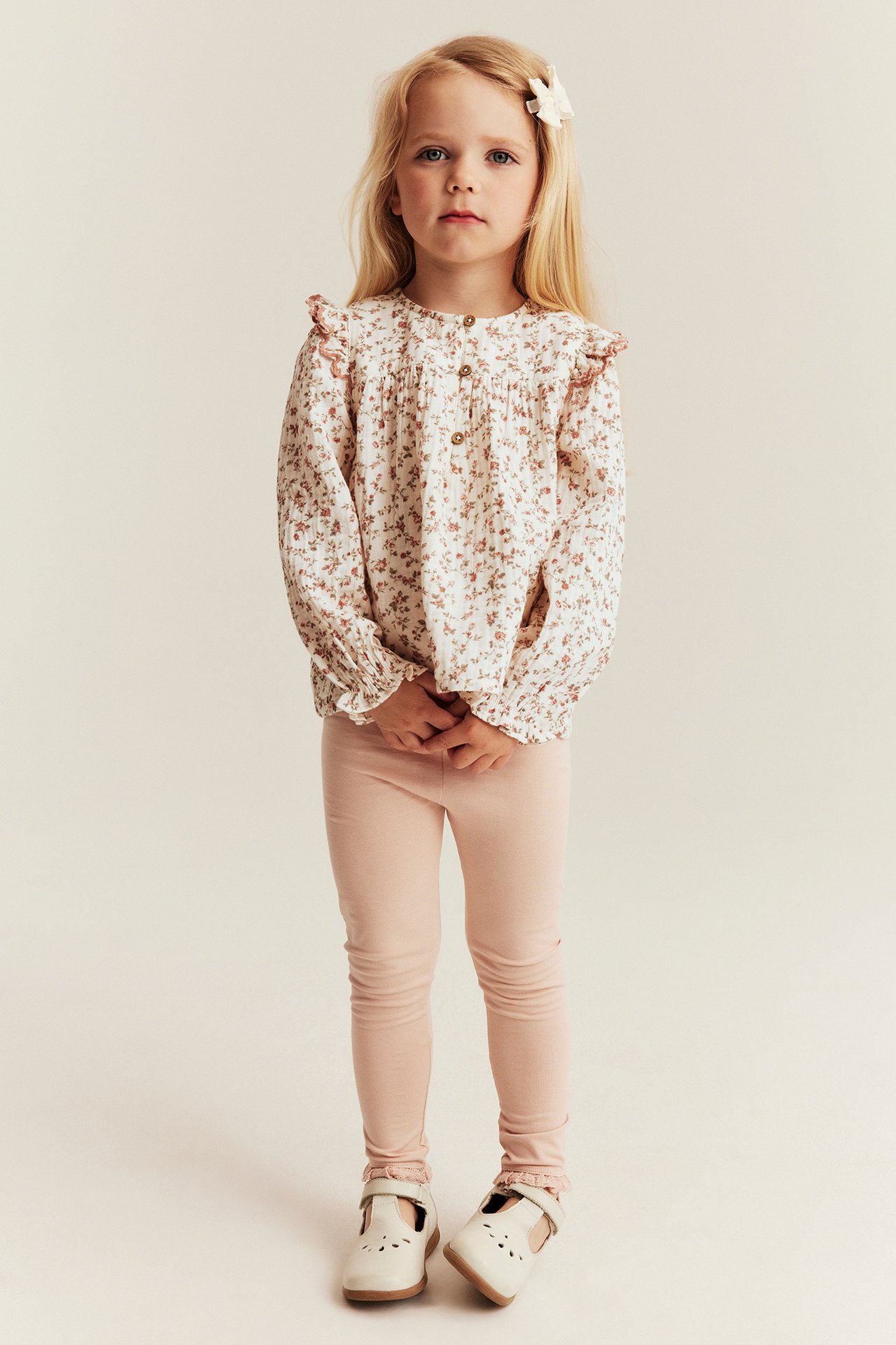 Forfra: Barn i offwhite blomstret musselinbluse, lange ermer, rosa leggings og hvite sko.