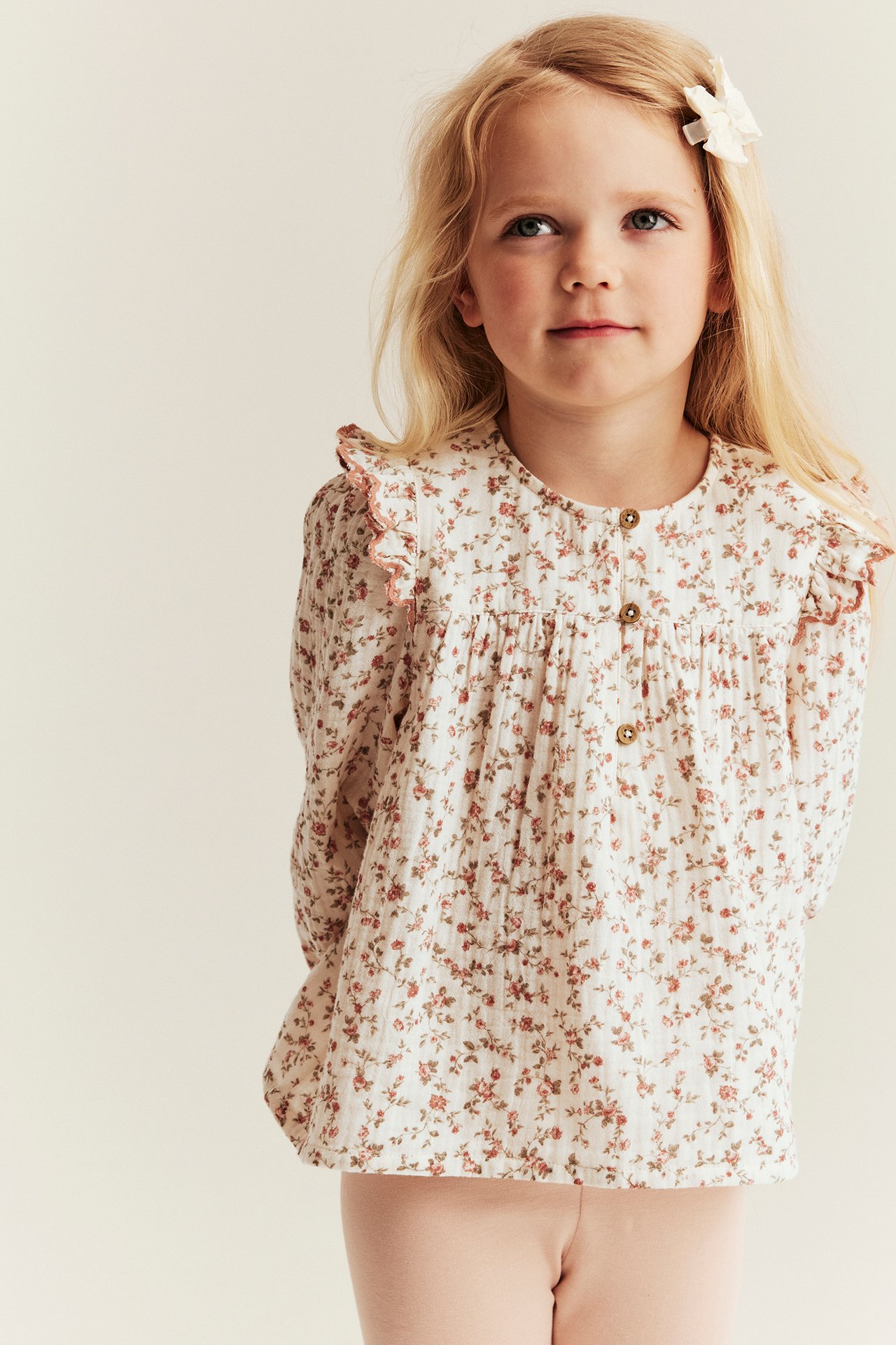Front: barn i offwhite blomstret muslinbluse med skulderkapper og lyserosa bukse.