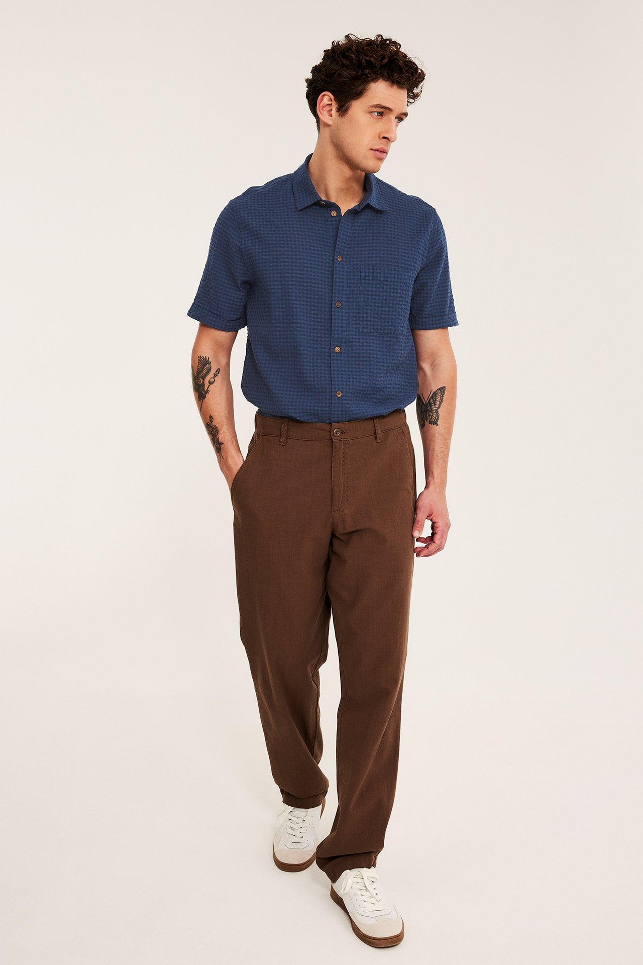 Framifrån man i blå kortärmad skjorta med bäckeböljetextur, bruna byxor och vita beiga sneakers.