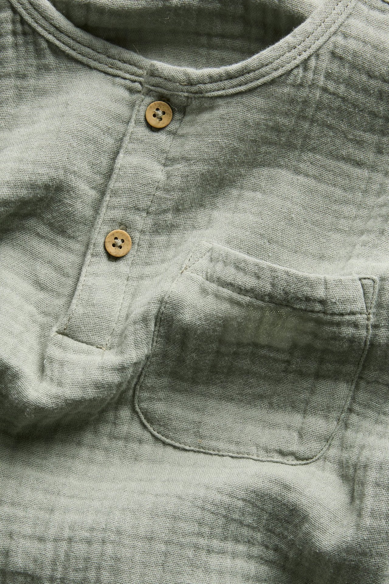 Närbild framifrån av en ljusgrön långärmad topp i bomullsmuslin med knappslå och bröstficka.