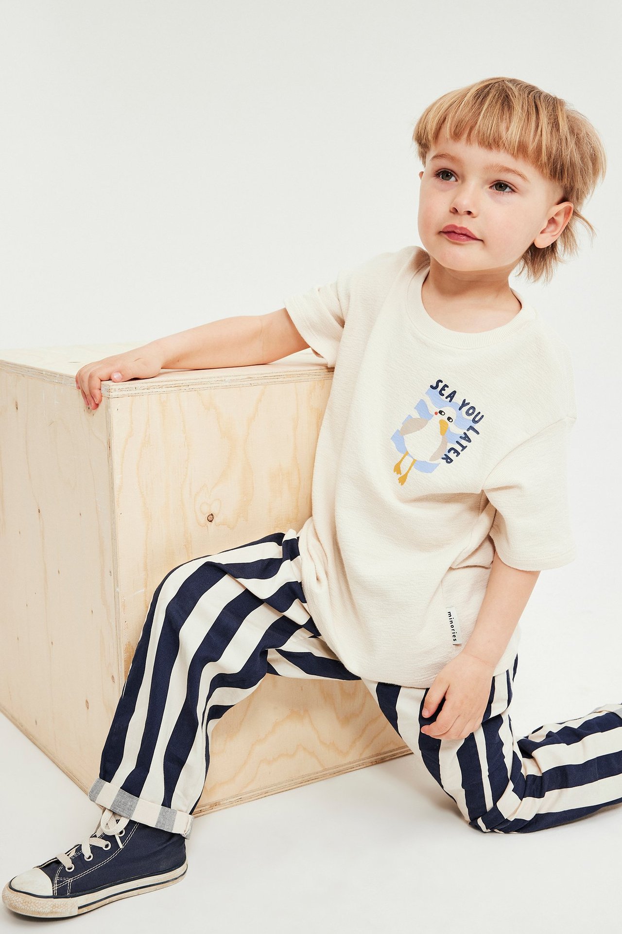 Från framsidan: barn i blå/benvita randiga bomullsbyxor, benvit t-shirt med måstryck och blå sneakers.