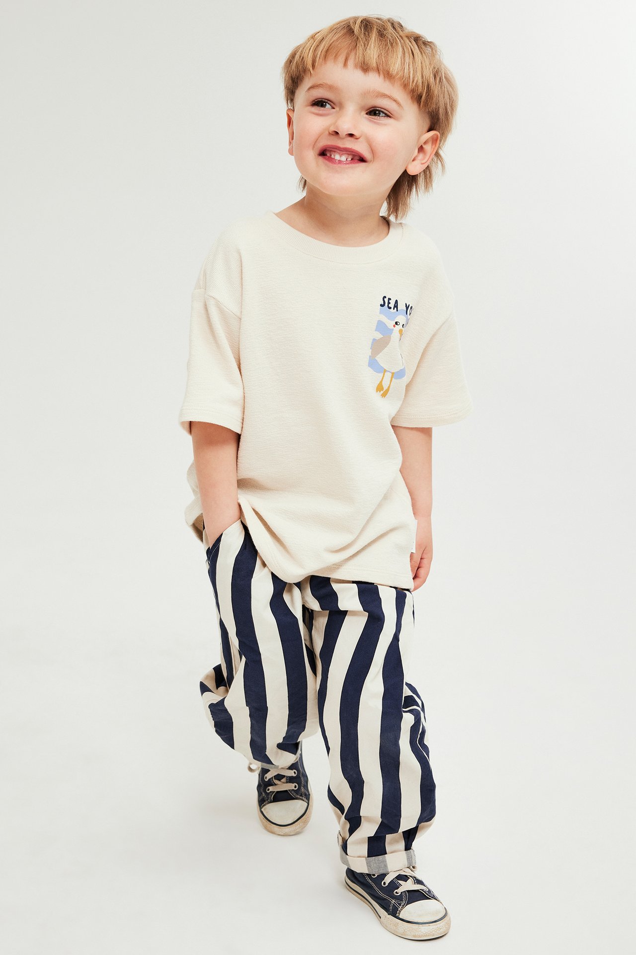 Frontvy av ett leende barn i blårandiga bomullsbyxor med uppvikta benslut och krämfärgad T-shirt.