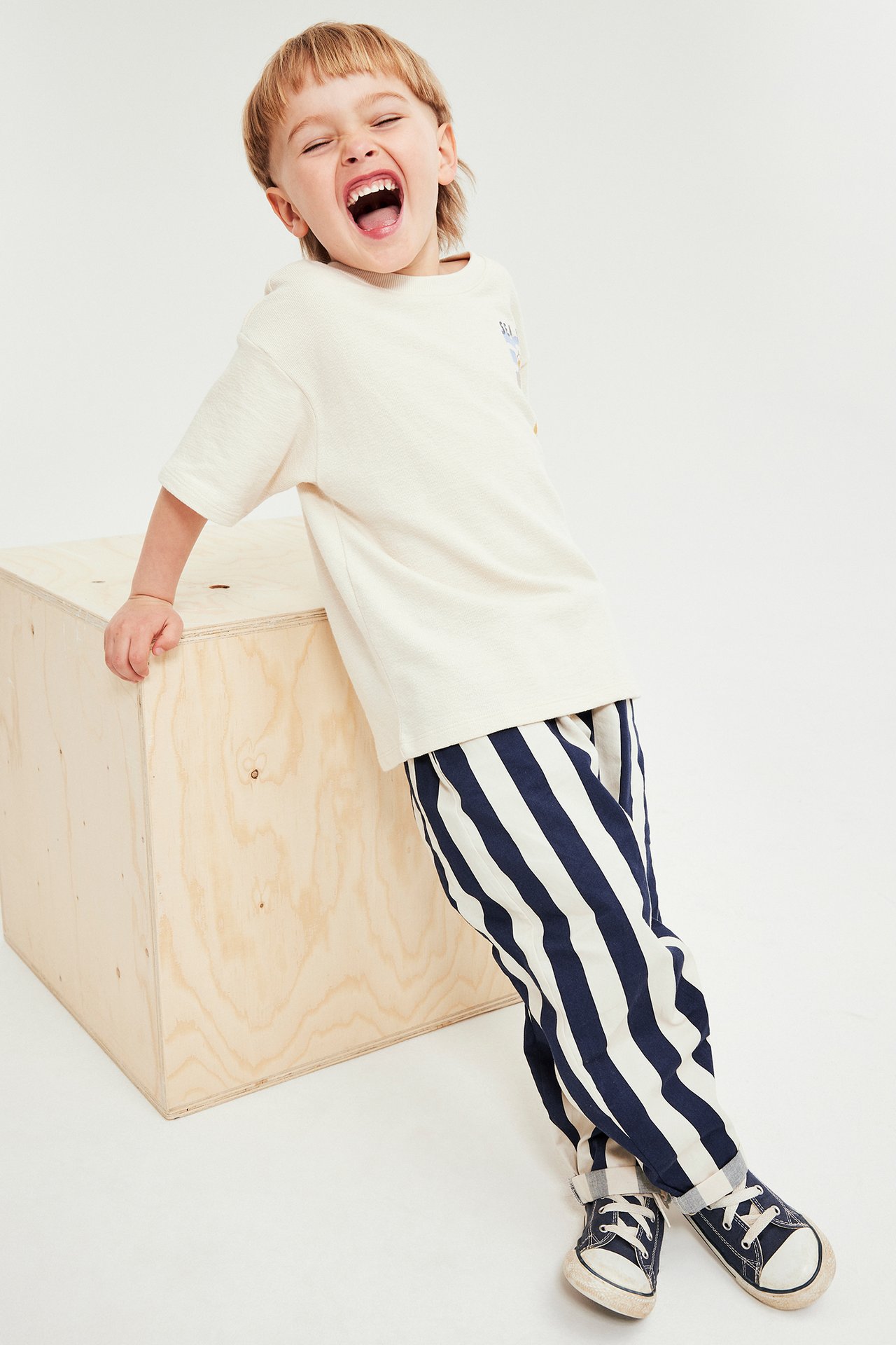 Edestäpäin naurava lapsi yllään sini-valkoraitaiset kudotut puuvillahousut, kermanvärinen t-paita ja tummansiniset lenkkarit.
