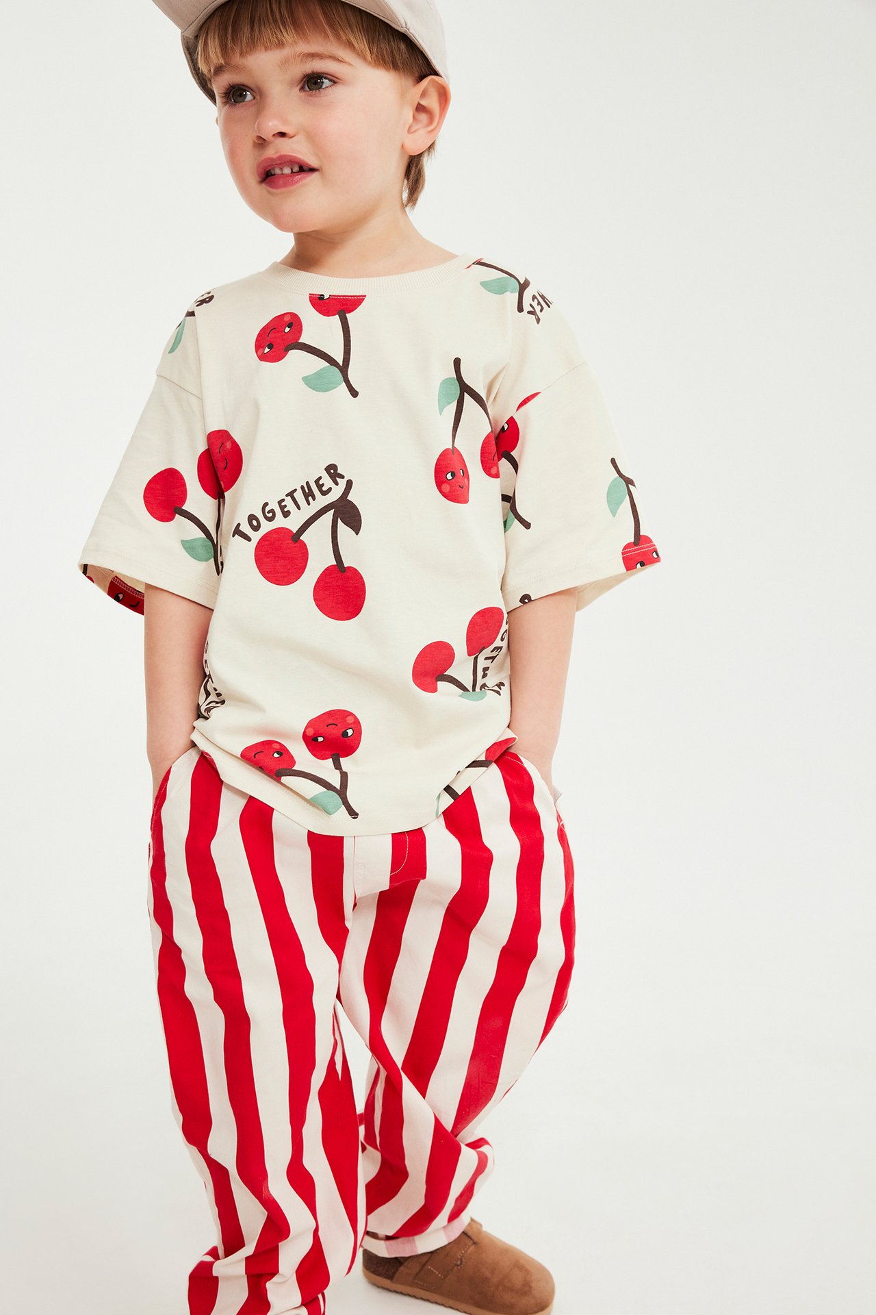 Edestä: Lapsi punavalkoraidallisissa puuvillahousuissa, kirsikkakuvioisessa paidassa ja beigessä lippalakissa.