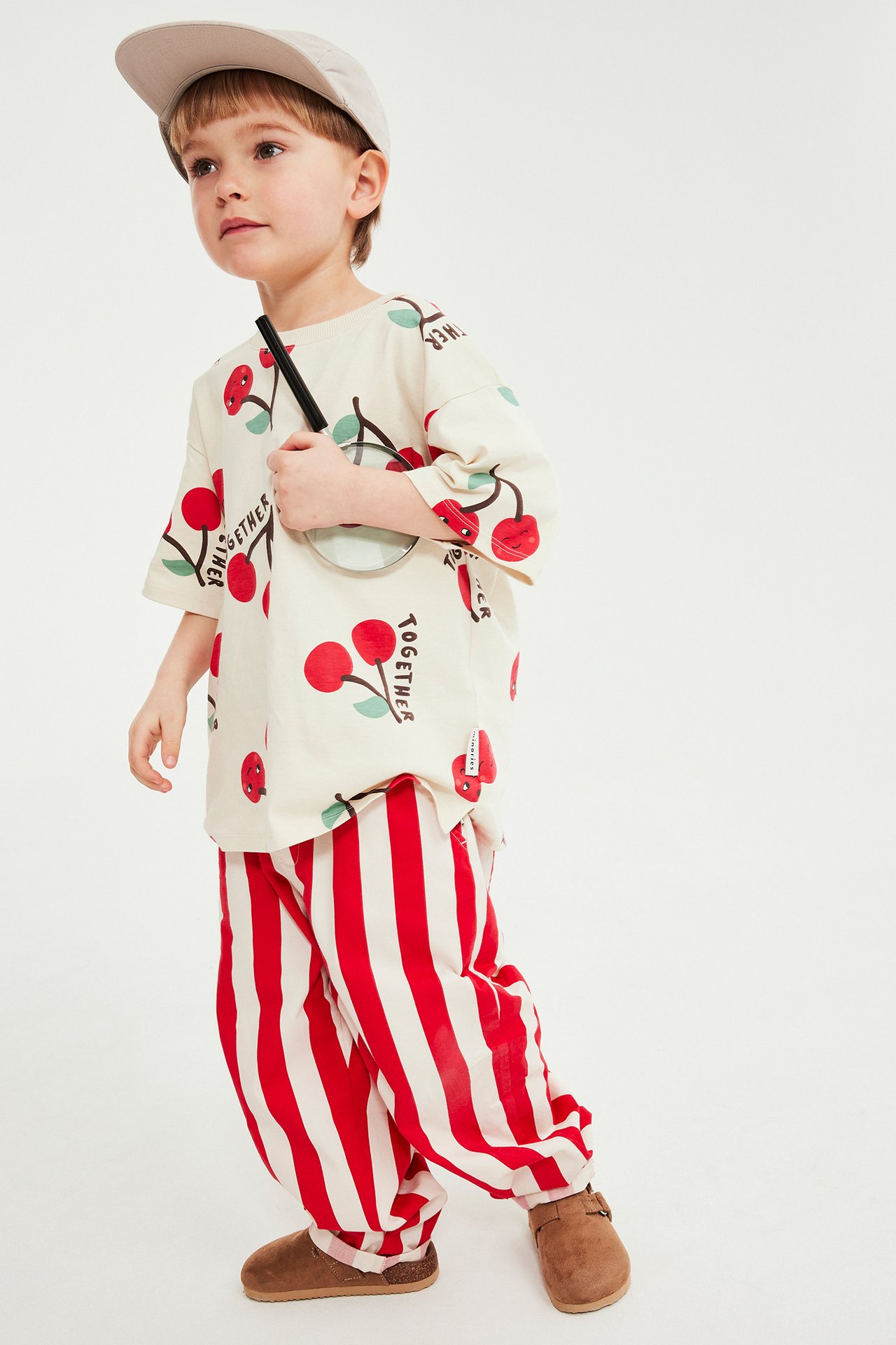 Framifrån: Barn i röda randiga bomullsbyxor, krämfärgad körsbärst-shirt, beige keps och bruna träskor.