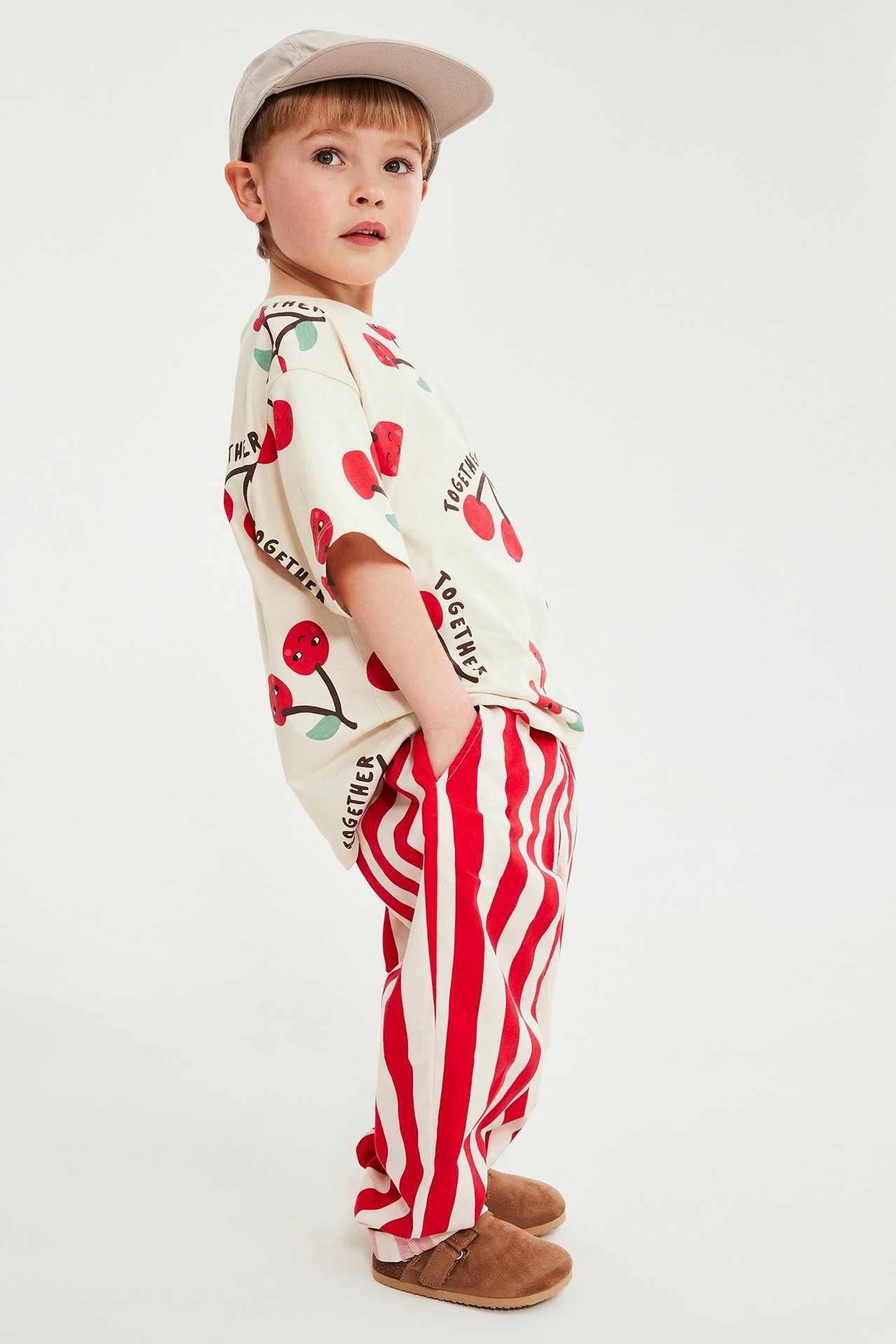Sidovy av barn i röd-vit randiga bomullsbyxor med fickor, körsbärsmönstrad t-shirt och beige keps.
