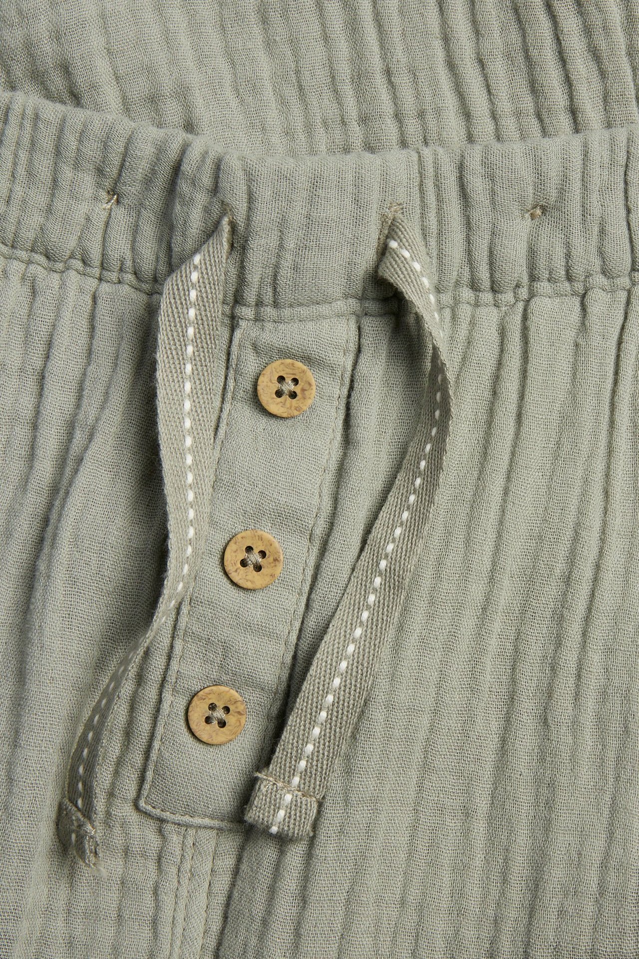 Zbliżenie ciemnozielonych spodni z muślinu bawełnianego z elastyczną talią, sznurkiem w jodełkę i ozdobnymi drewnianymi guzikami.