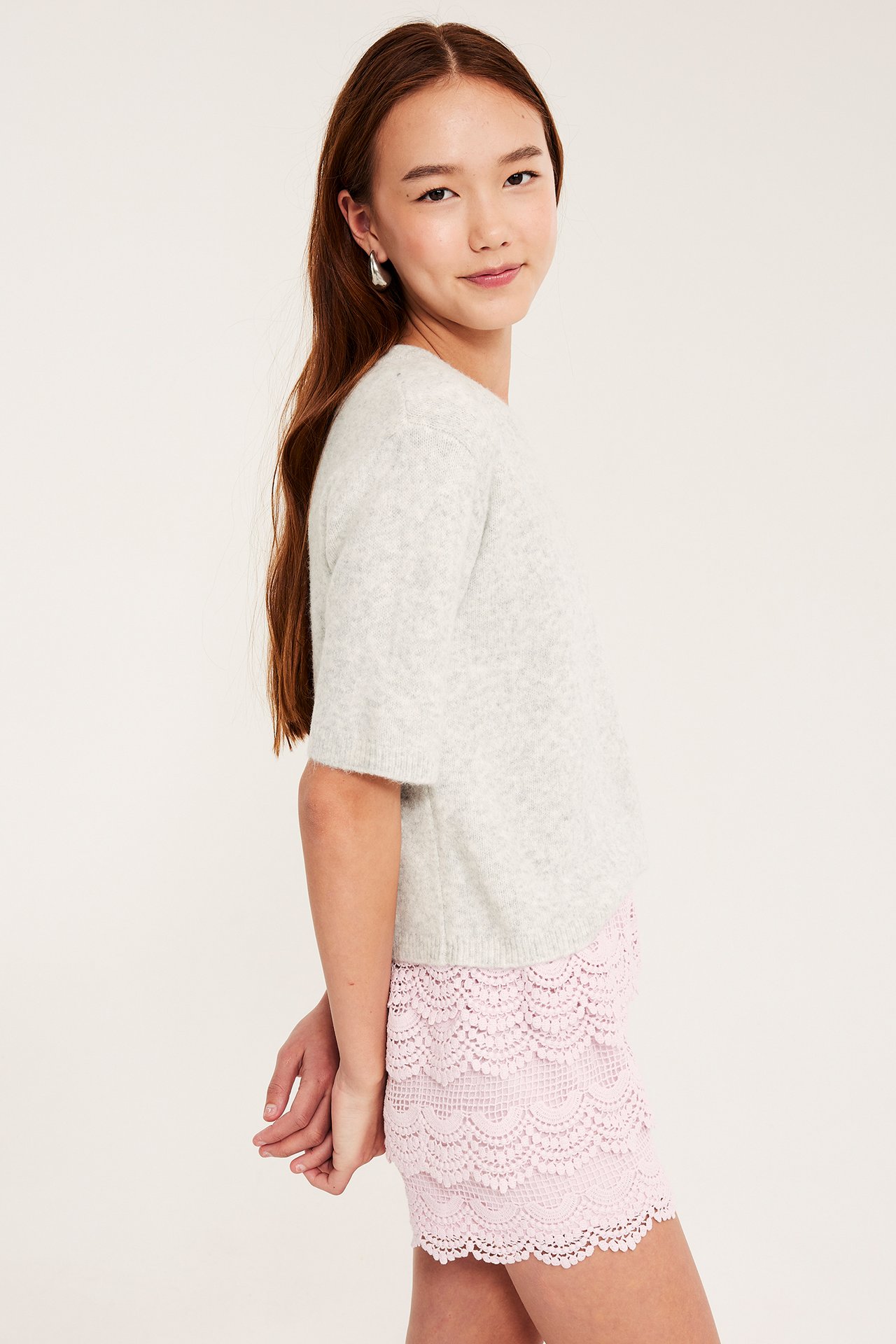 Widok z boku: Młoda kobieta w jasnoróżowych, warstwowych koronkowych szortach i jasnoszarym swetrze z krótkim rękawem.