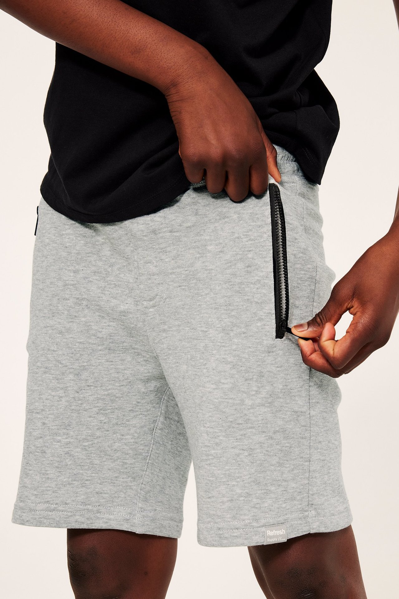 Edestäpäin: lapsi yllään vaaleanharmaameleeratut college-shortsit mustilla vetoketjutaskuilla. Käsi avaa vetoketjua.