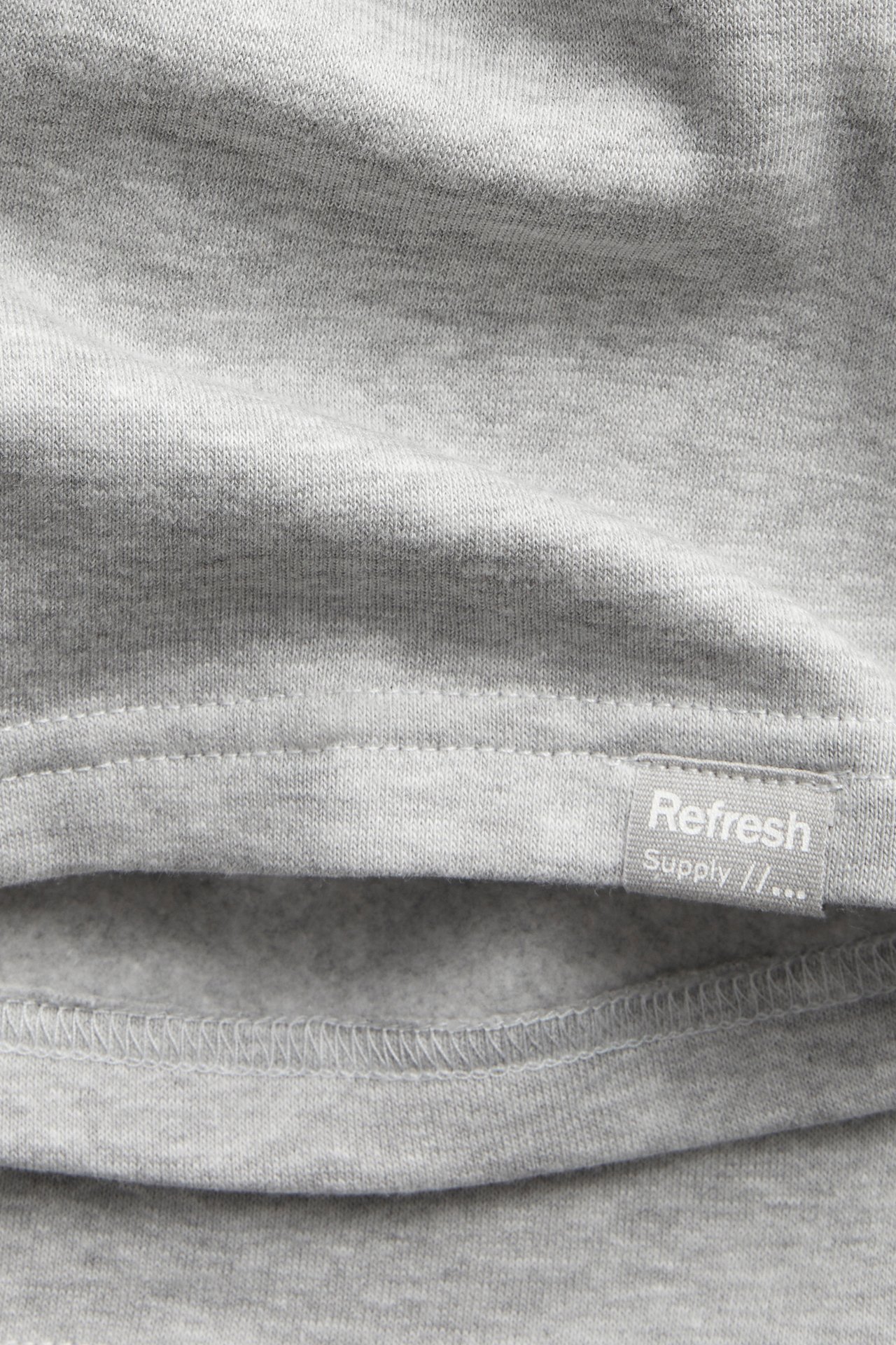 Närbild av ljusgrå melerad bomullstrikå från sweatshorts med liten "Refresh Supply"-etikett.