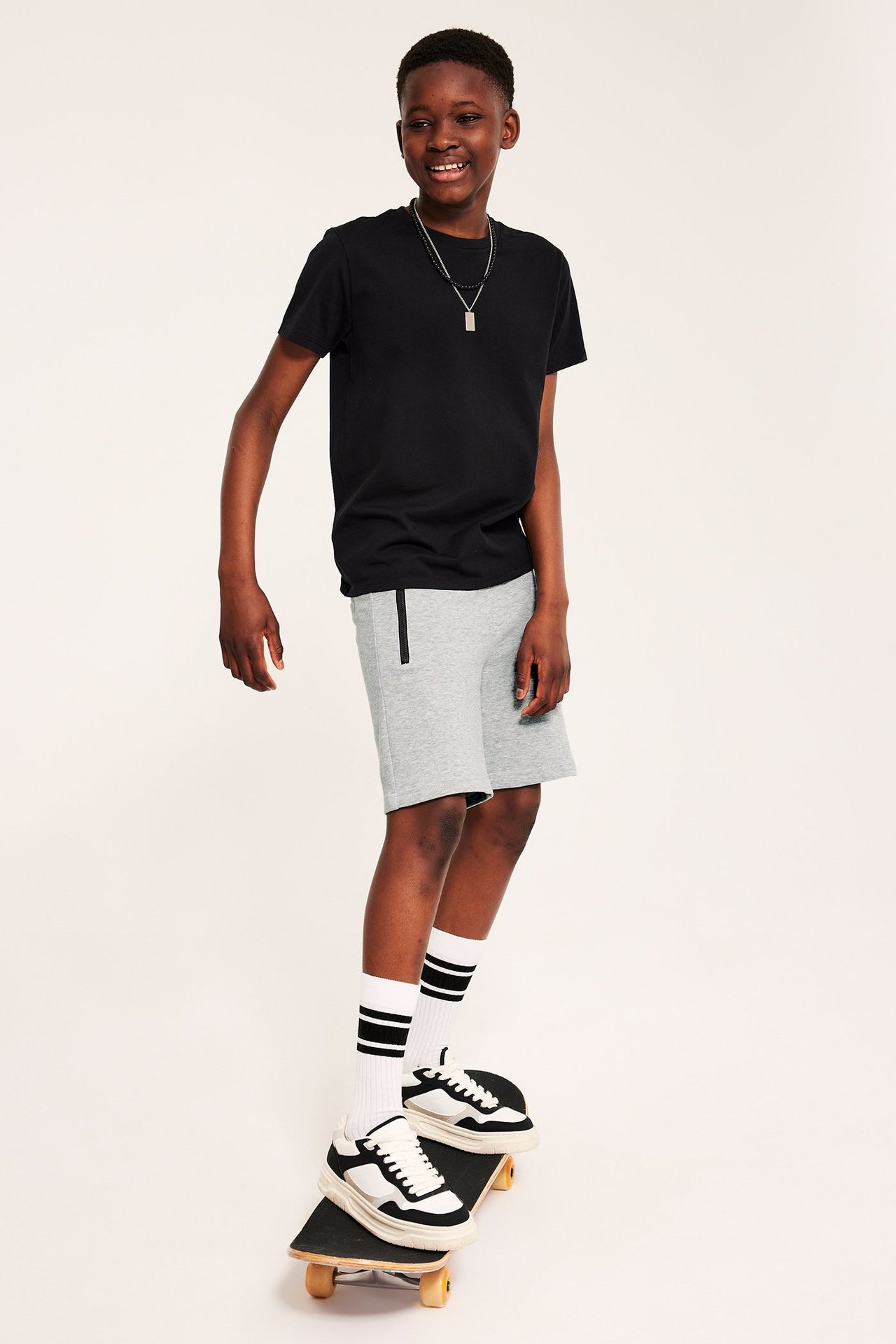 Edestä kuvattuna lapsi yllään vaaleanharmaamelange-collegehousut mustilla vetoketjuilla, musta t-paita ja lenkkarit.