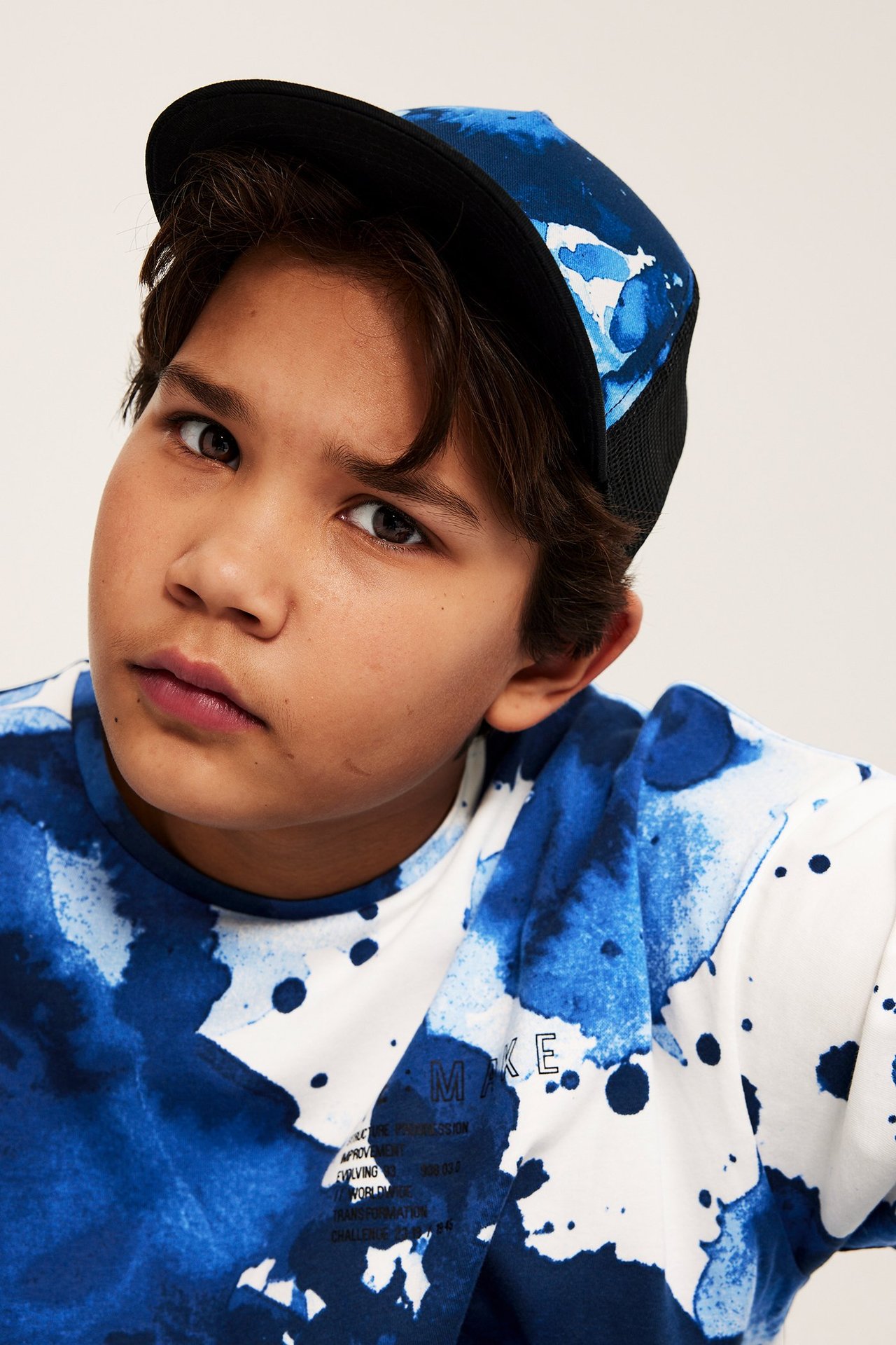 Widok z przodu dziecka w niebieskiej wzorzystej czapce tirówce z czarnym daszkiem i pasującej koszulce.