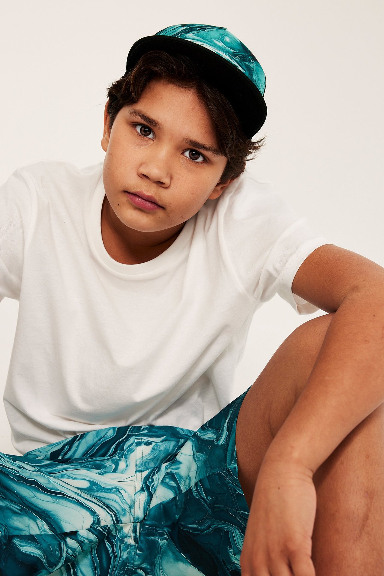 Framifrån, ett barn bär en grön marmorerad truckerkeps, en vit t-shirt och matchande gröna mönstrade shorts.