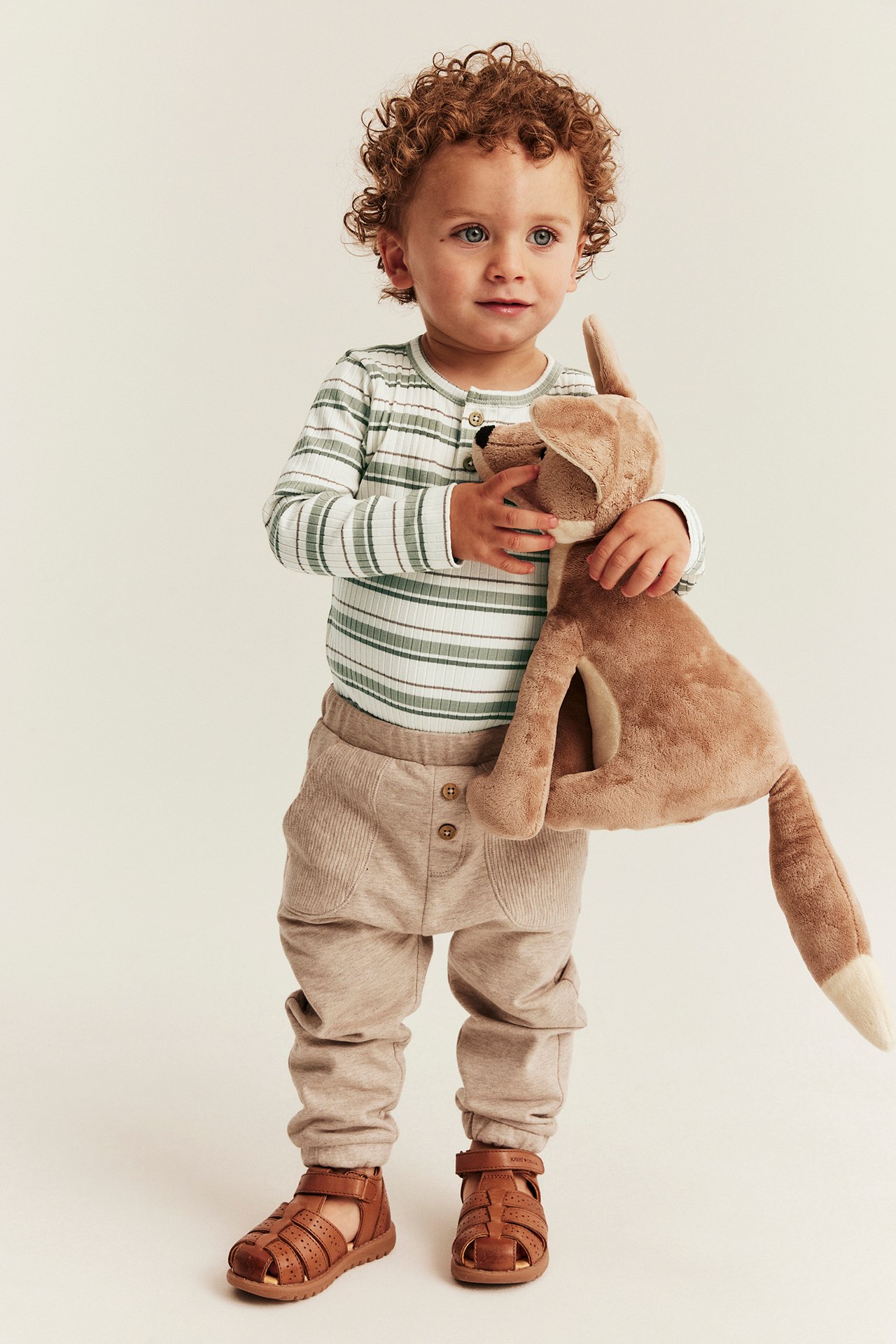 Forfra: babygutt i ribbet hvit/grønnstripet langermet body, beige bukser og brune sandaler, holder en kose-rev.