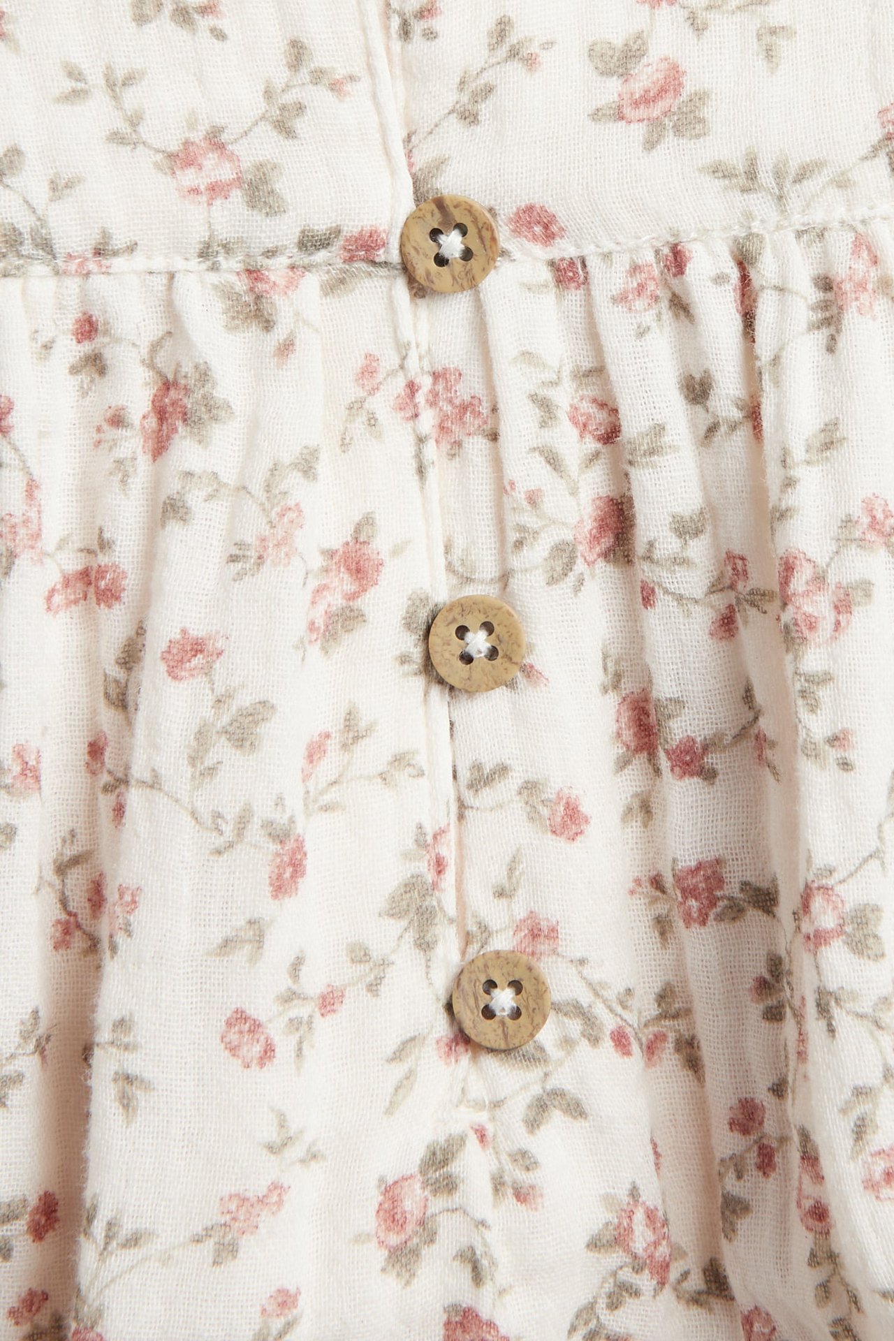 Närbild av en off-white babybody i bomullsmuslin med småblommigt mönster och tre träknappar.