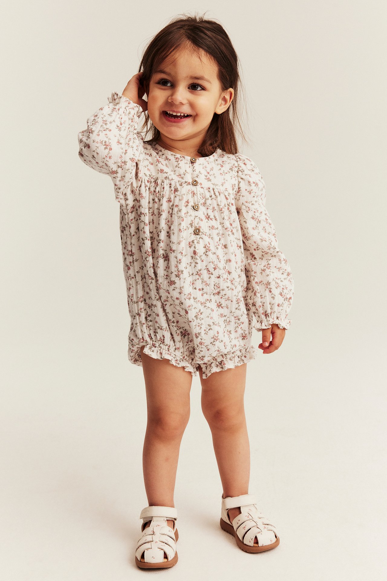 Frontalt bilde av et smilende barn i offwhite blomstret musselinbody med lange ermer og lysbeige sandaler.