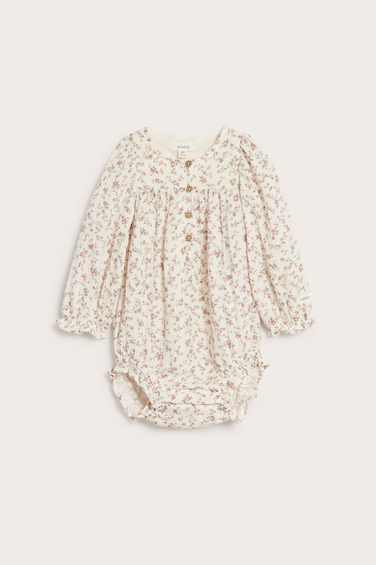 Framifrån, off-white baby långärmad muslinbody med smått blommigt mönster, resårmanschetter och knappar framtill.