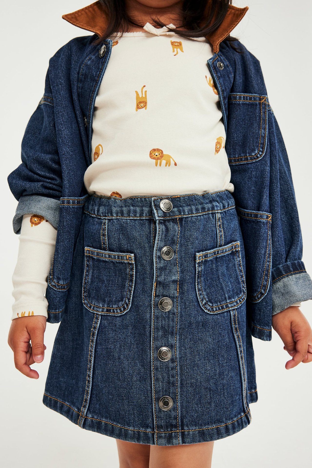 Front: Barn i mørkeblå knappedenimskjørt, topp med løvetrykk og åpen denimjakke.