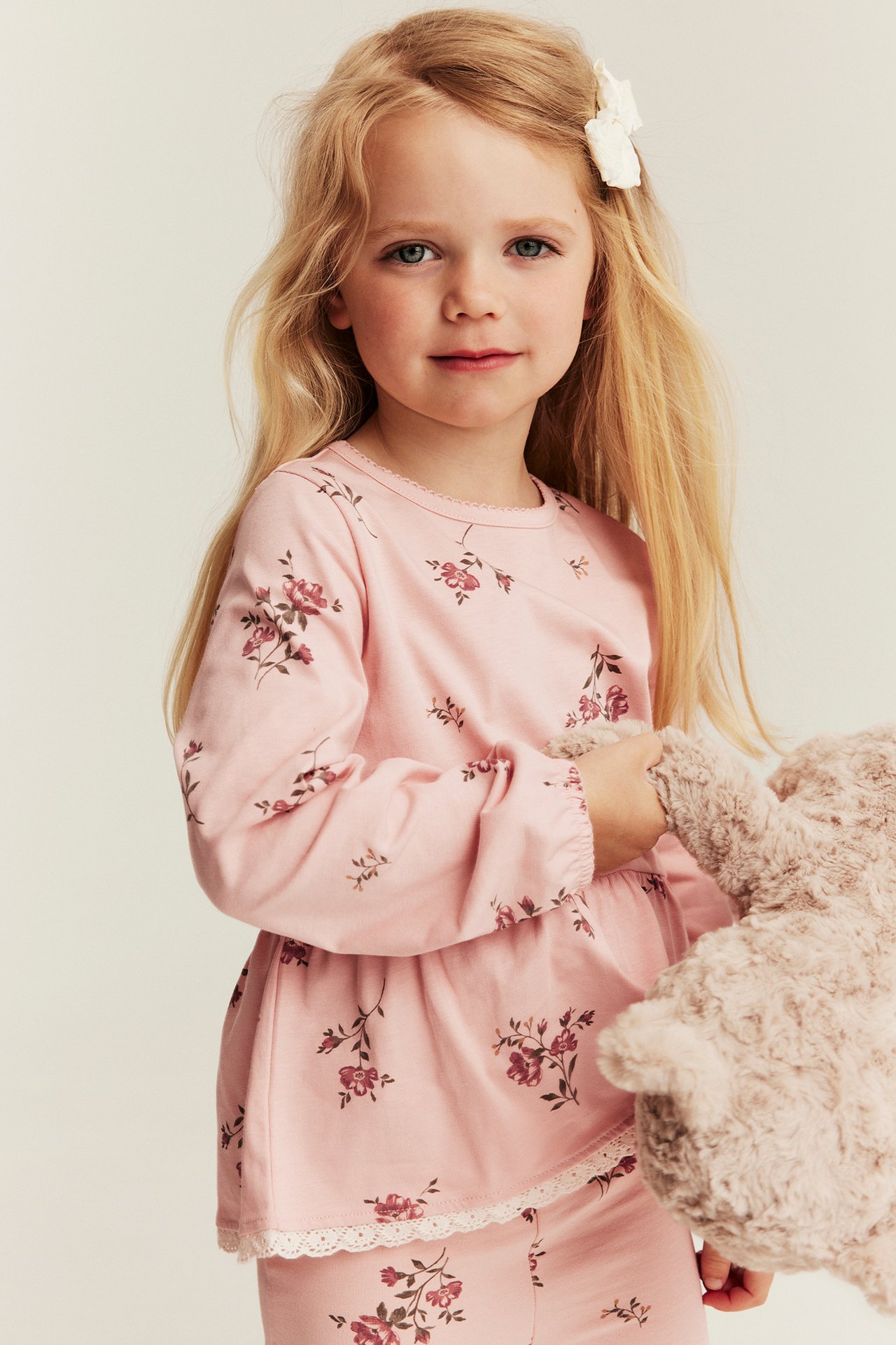 Frontvisning, barn i rosa blomstret peplumtopp med blondekant og picothals, holder en bamse.