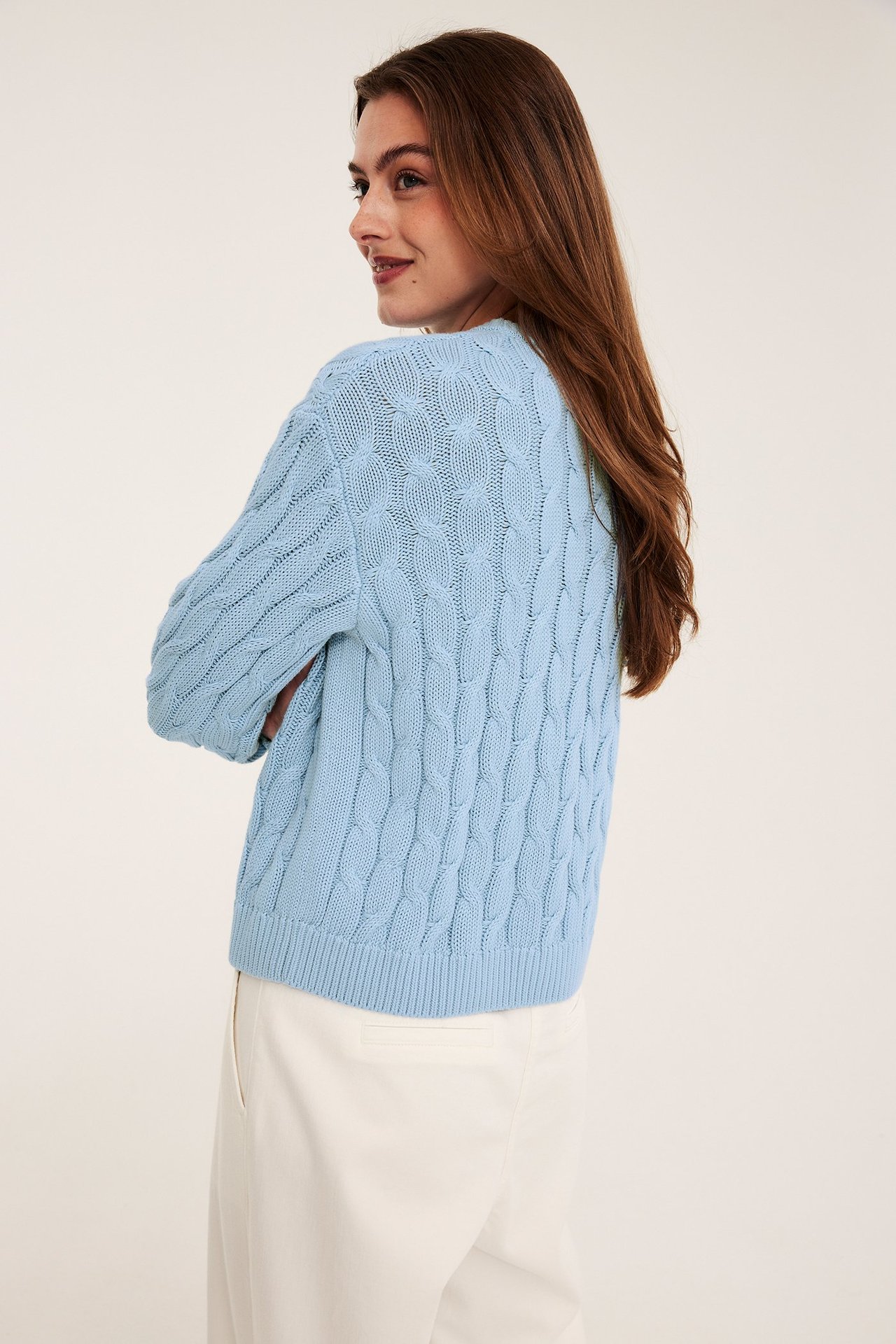 Sidovy bakifrån av en kvinna i ljusblå kabelstickad bomullströja över vita byxor.