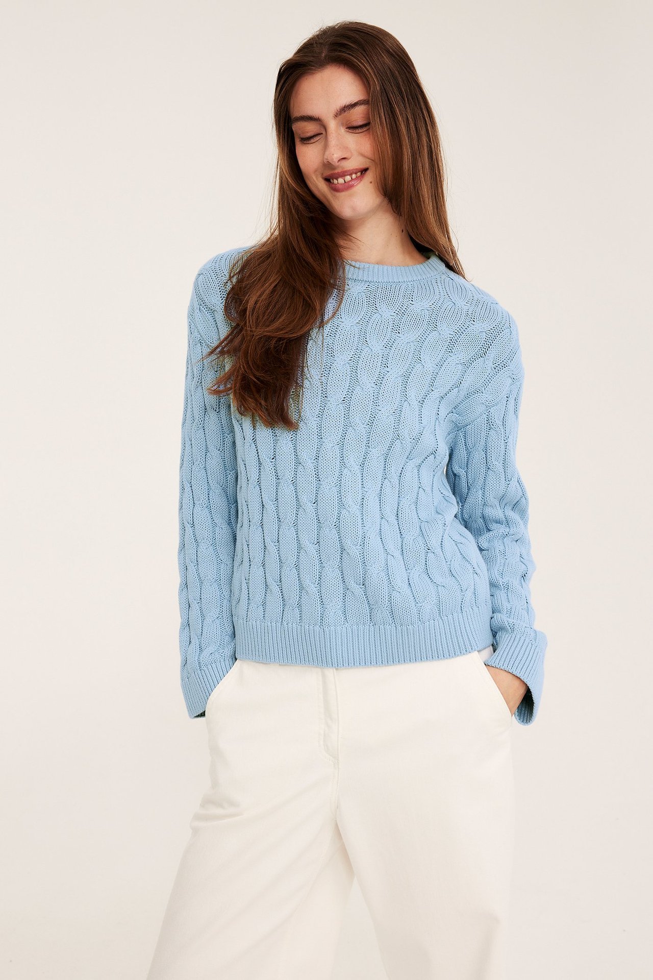 Edestäpäin nainen yllään vaaleansininen palmikkoneulottu puuvillapusero ja valkoiset housut.