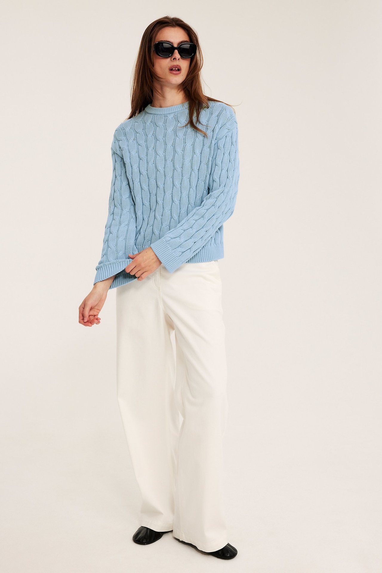 Nainen vaaleansinisessä palmikkoneuleessa, valkoisissa leveissä housuissa, mustissa aurinkolaseissa ja mustissa kengissä.