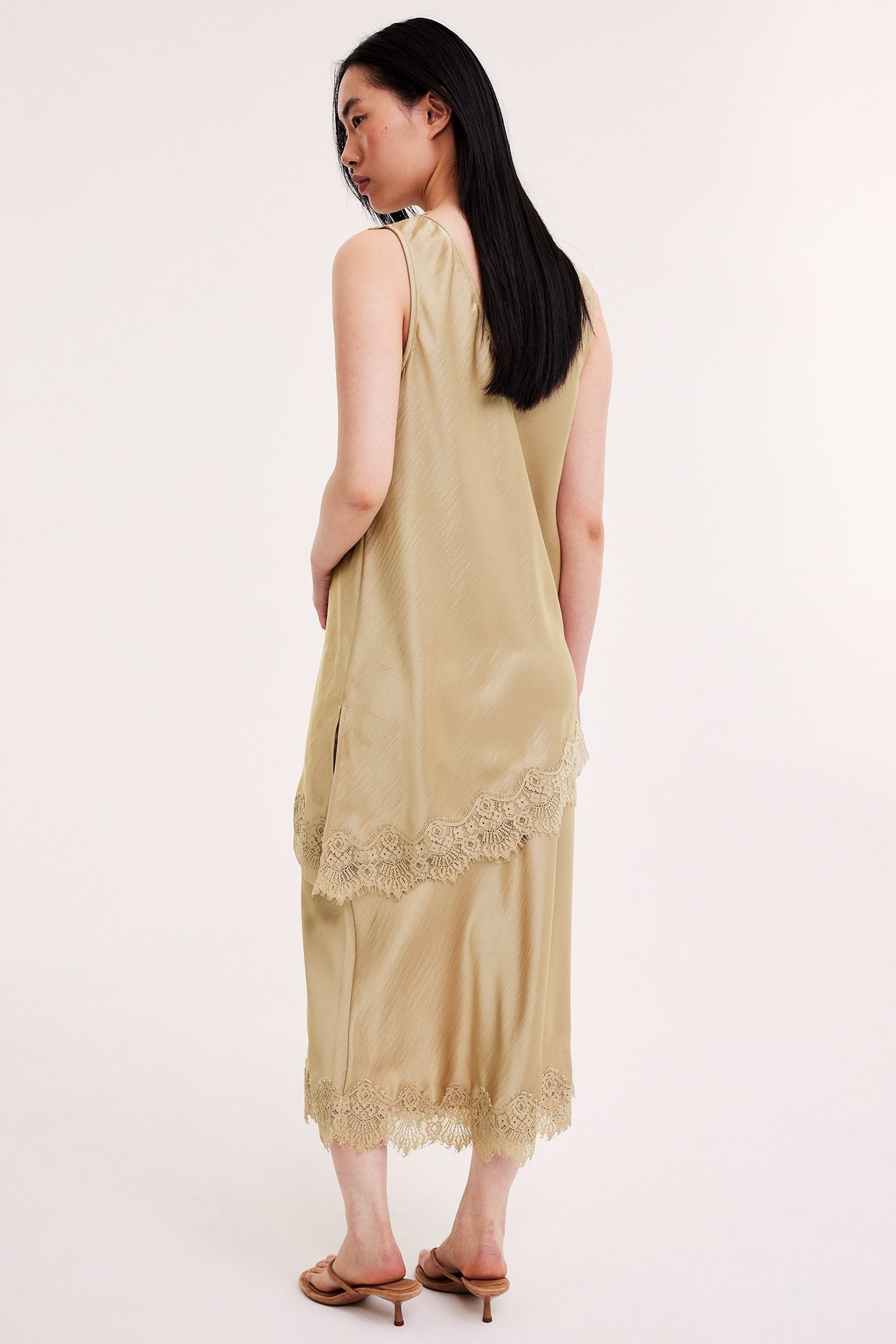 Bak-sidevisning av kvinne i lys brun sateng slipkjole med blonder og matchende sandaler med hæl.