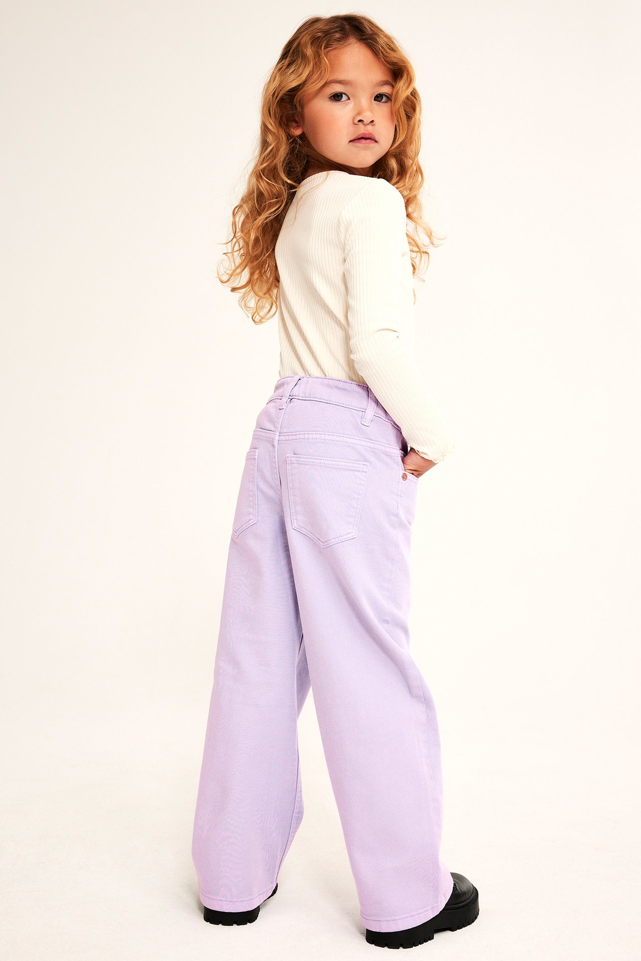 Barn i lilla vide jeans, kremfarget ribbet langermet topp, svarte chunky sko, bakfra.