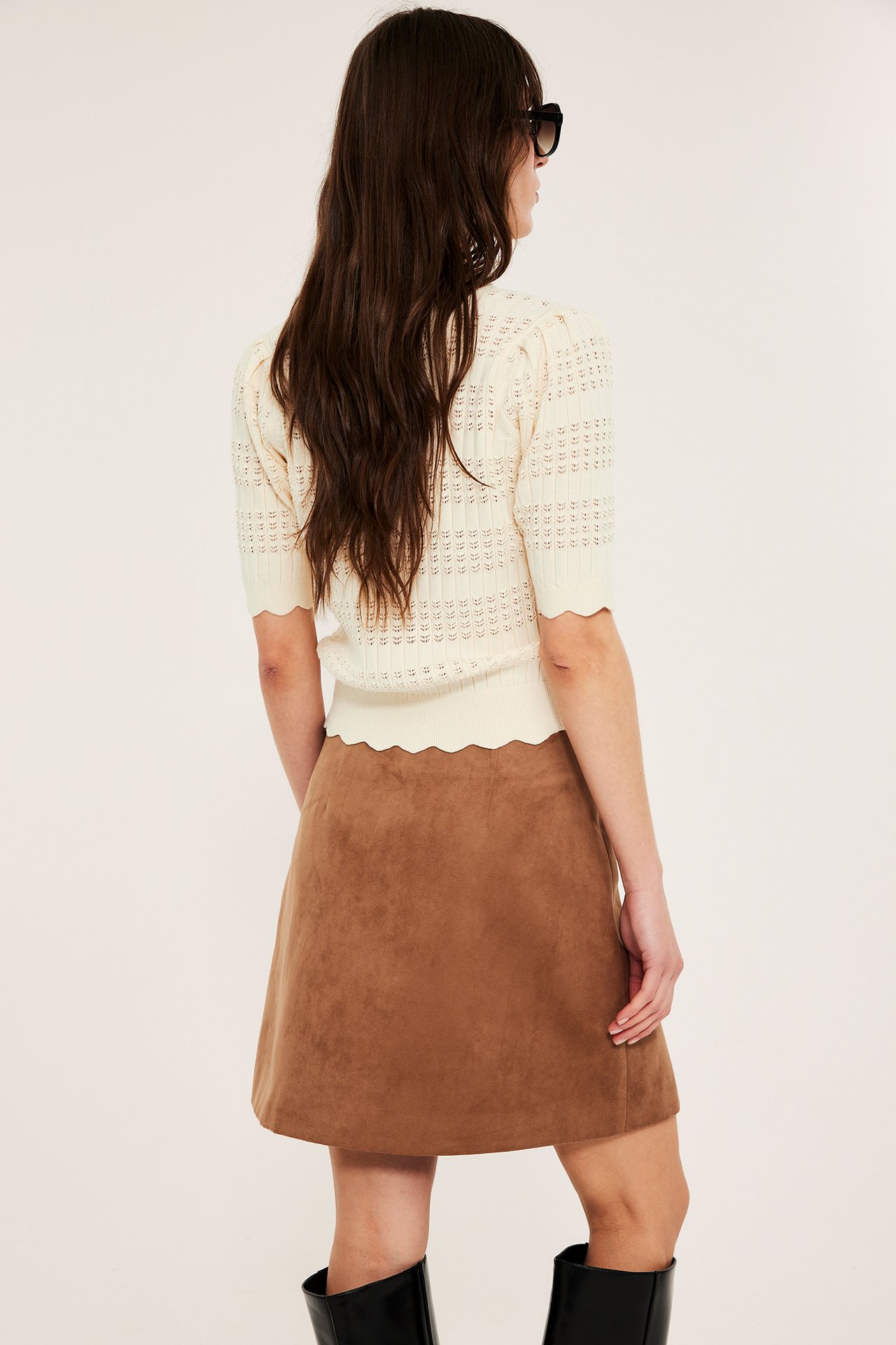 Widok z tyłu: kobieta w kremowym krótkim ażurowym topie z falistym brzegiem, brązowej spódnicy i czarnych butach.