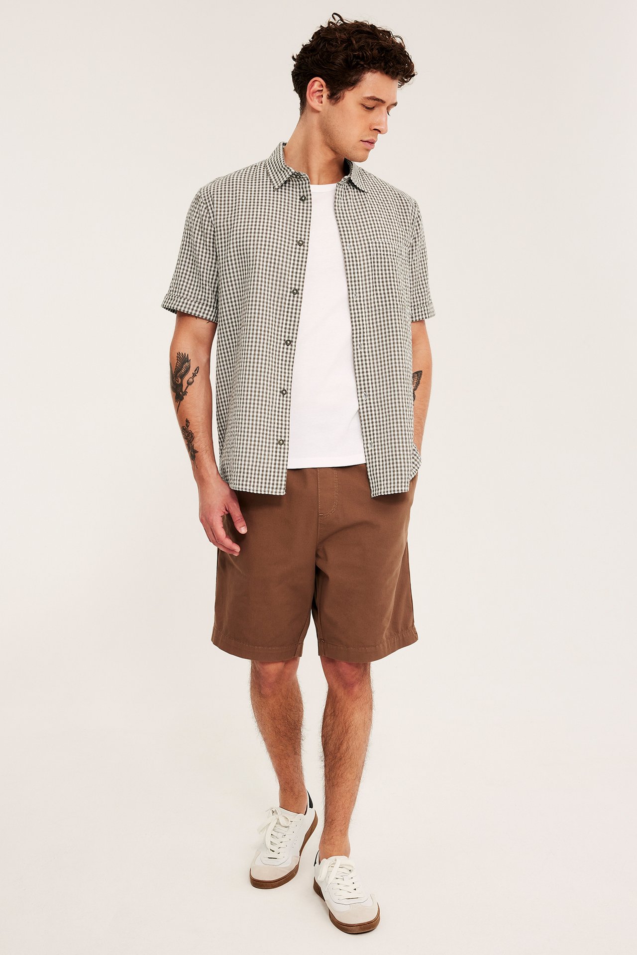 Frontbilde: Mann i lys brun rutete kortermet skjorte over hvit t-skjorte, brune shorts og hvite sneakers.