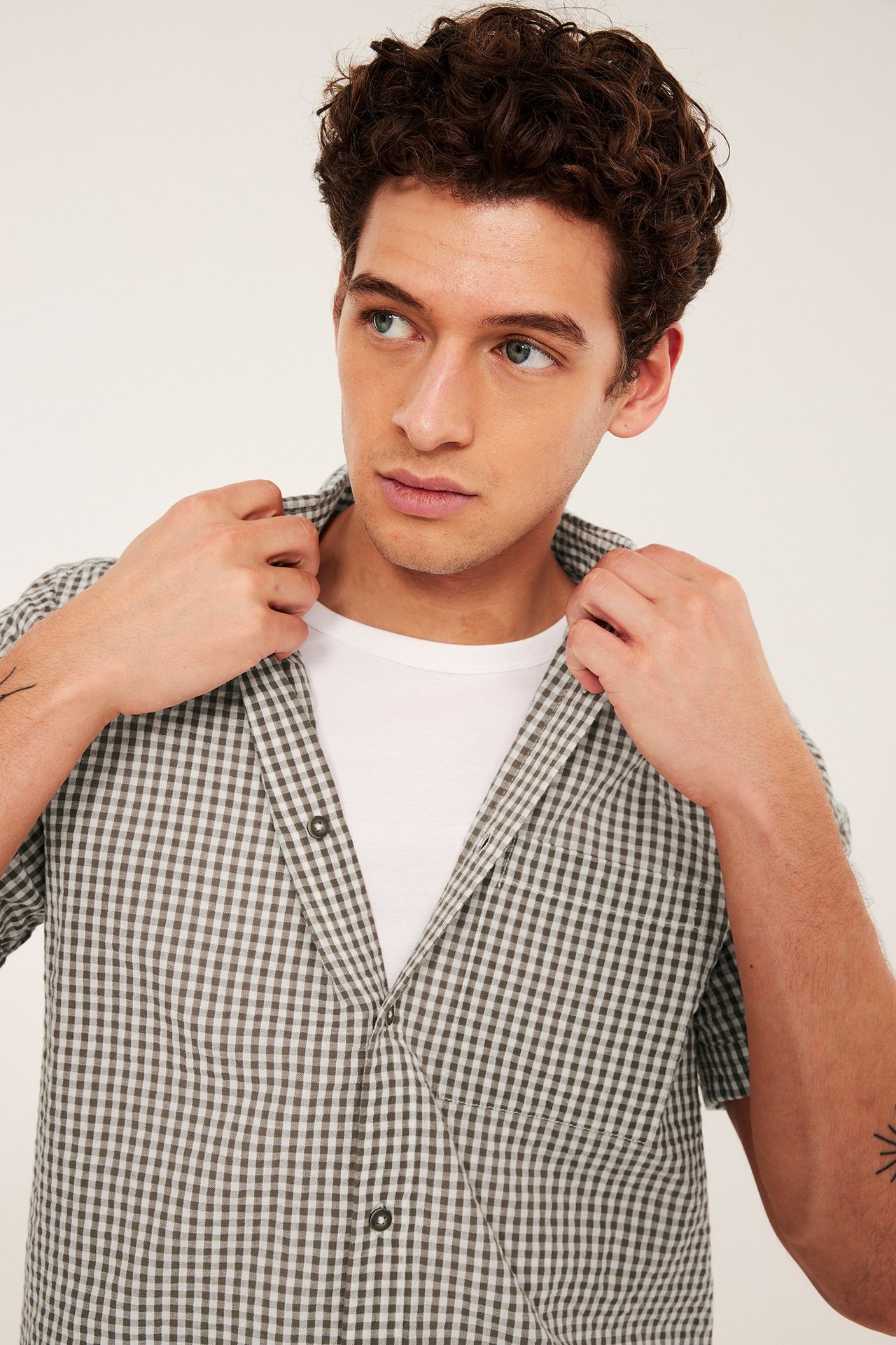 Närbild framifrån på en man i en öppen ljusbrunrutig kortärmad bäckeböljaskjorta över en vit t-shirt.