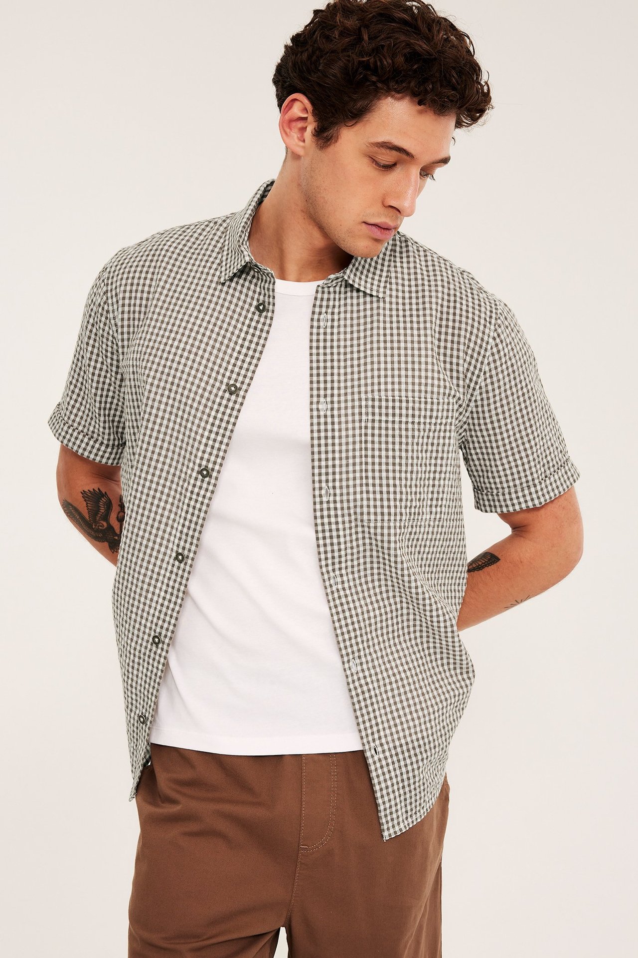 Frontvy av man i öppen ljusbrun rutig kortärmad bäckeböljaskjorta över vit t-shirt och bruna byxor.