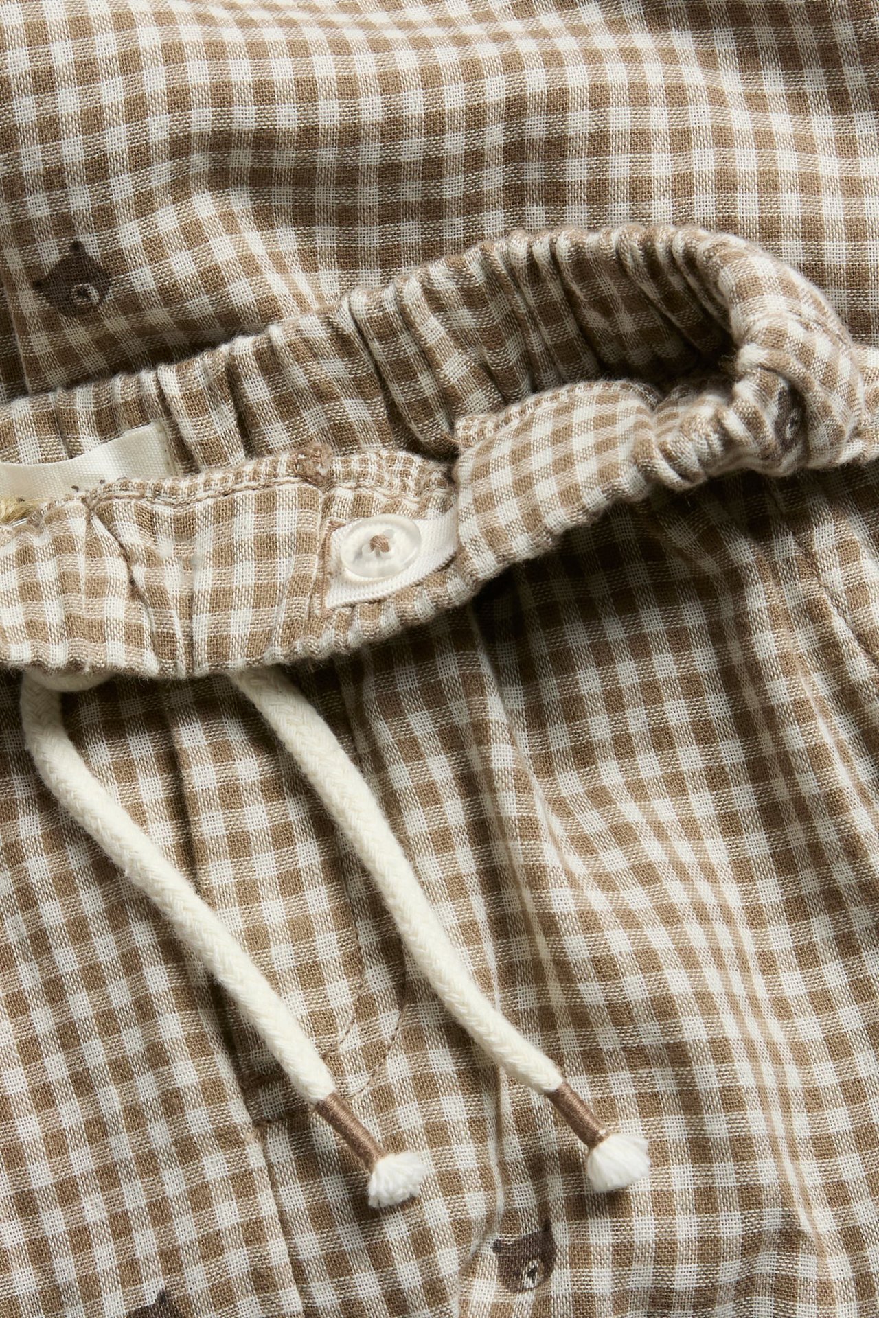 Nærbilde av brune og hvite rutete shorts med elastisk midje, knapp, snøring og bjørnetrykk.