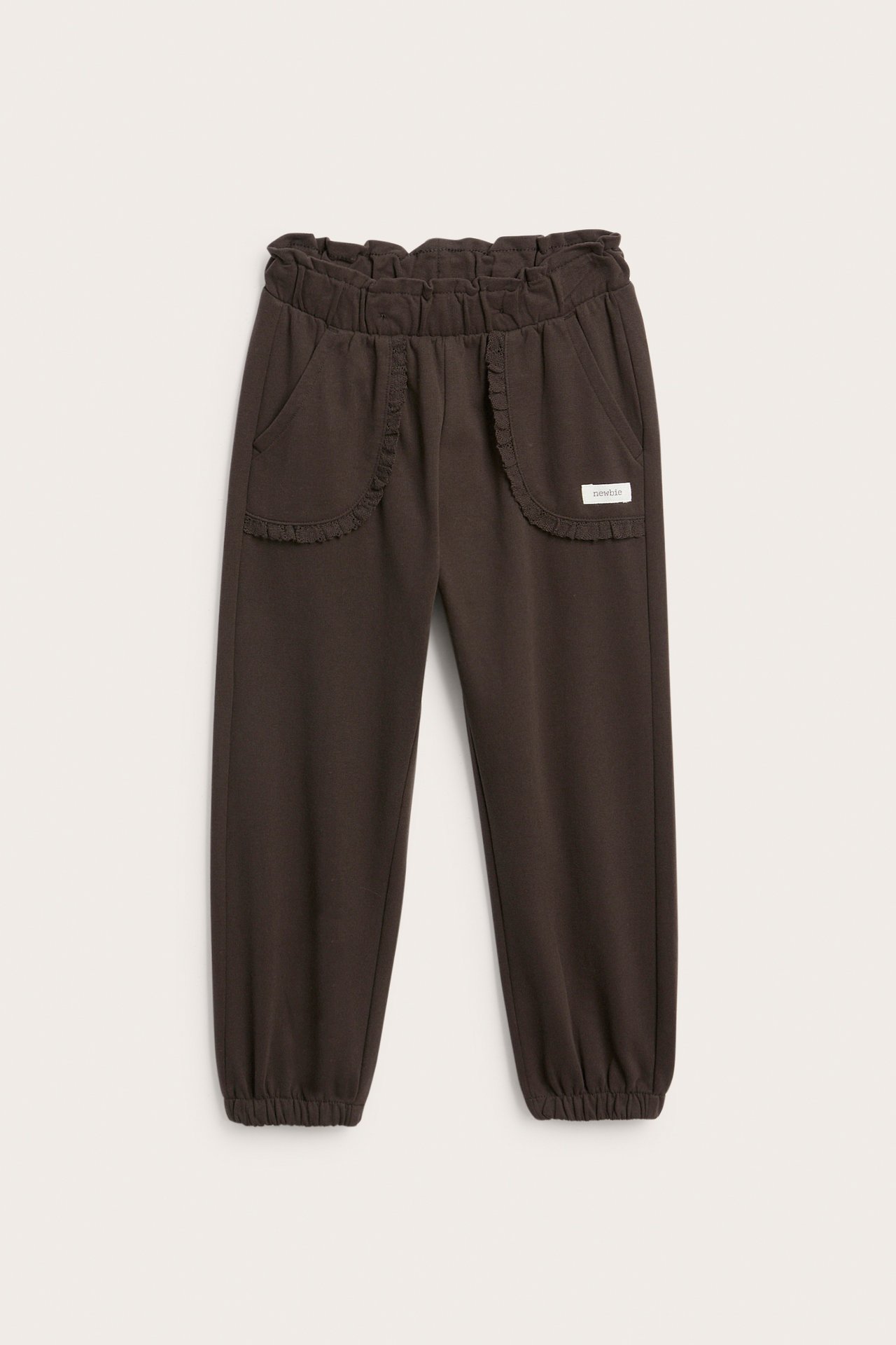 Frontvy av mörkbruna barnjoggers med volangmidja och fickor, samt resår i benslut.