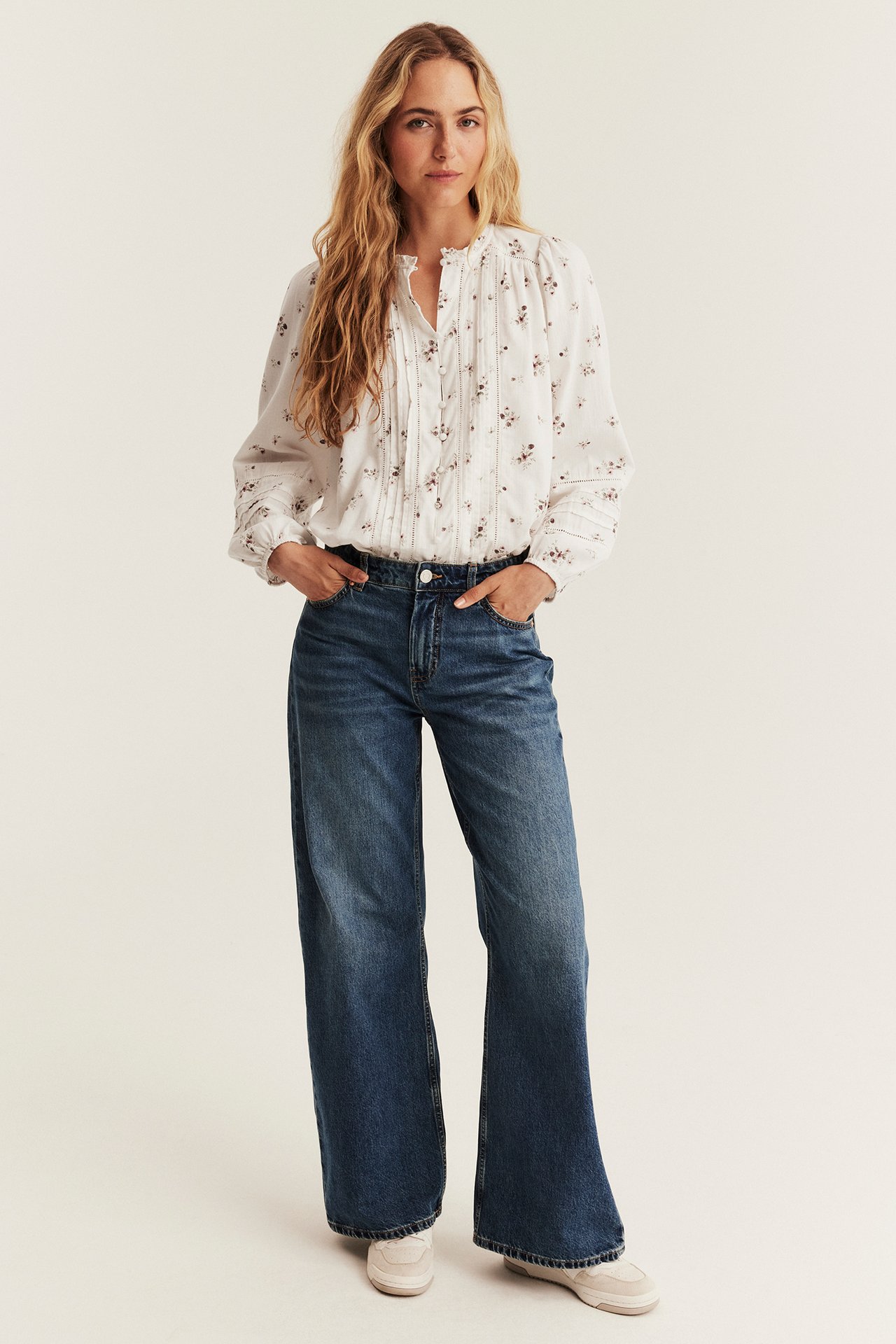 Front: kvinne i hvit blomstret folderbluse, blå vide jeans og hvite joggesko.