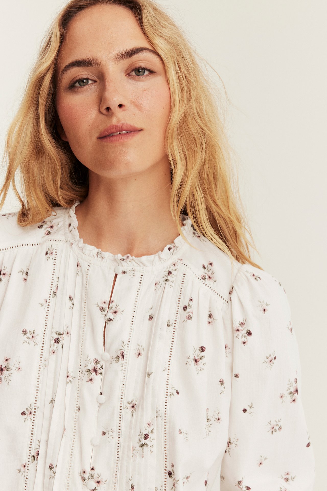 Nærbilde forfra av kvinne i hvit langermet blomstret bluse med folder og rysjekrage.