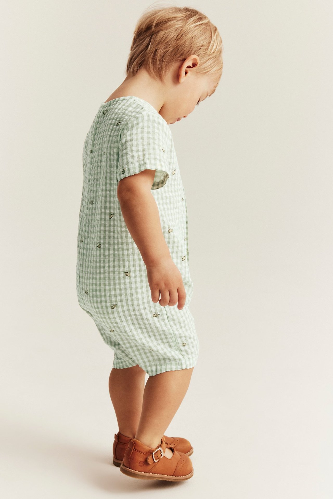 Sivukuva lapsesta vihreäruudullisessa mehiläiskuvioisessa puuvillaromperissa ja ruskeissa solkikengissä.