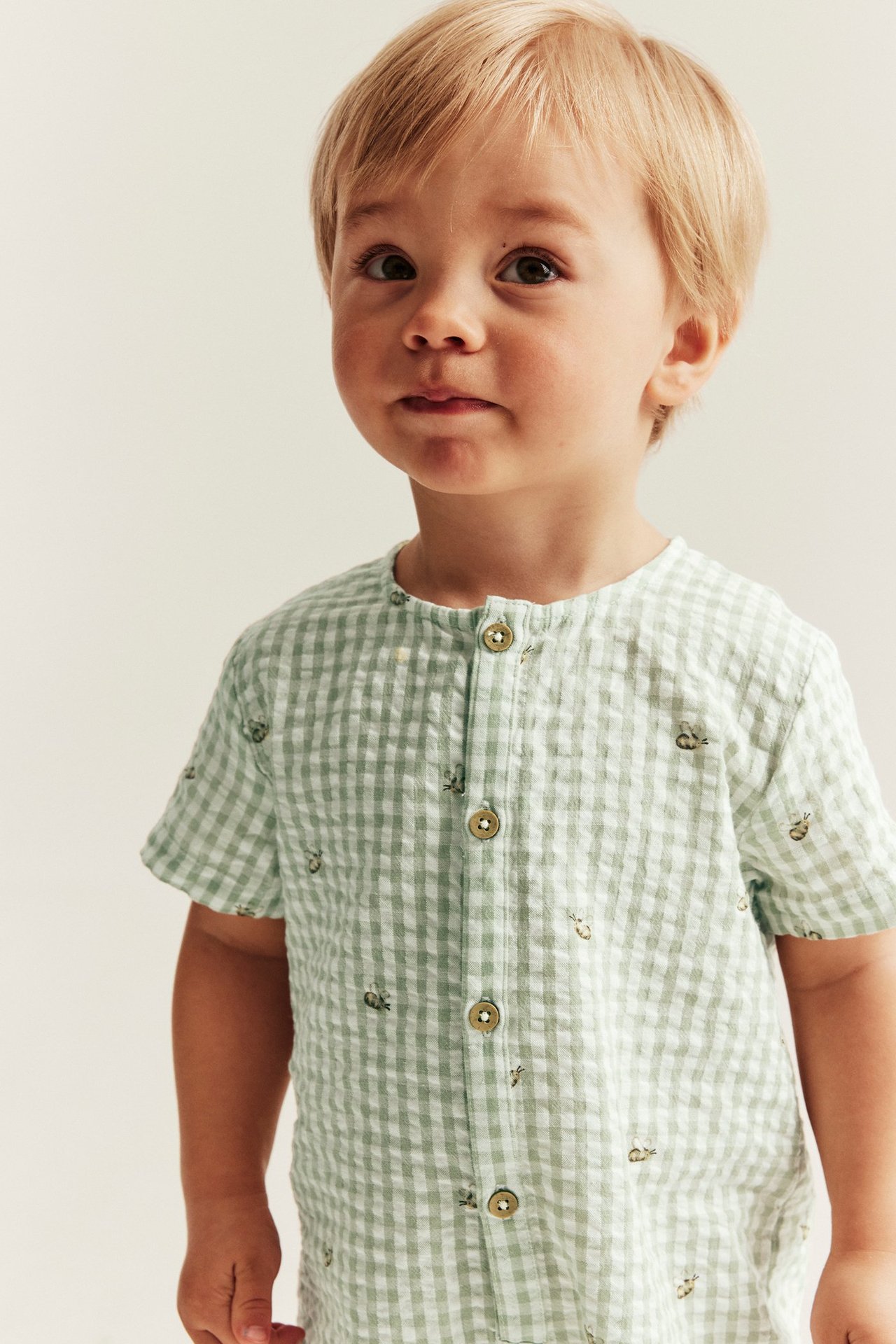 Frontvy av ett baby i en grön- och vitrutig bomullsjumpsuit med bi-mönster och knappar framtill.