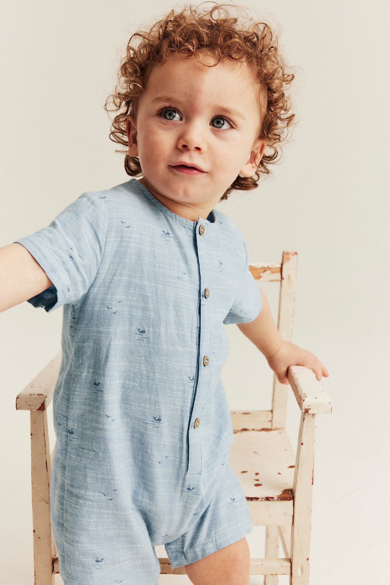 Foran-sidebilde av baby i lyseblå kortermet bomullsjumpsuit med hval- og fuglemønster, med knapper foran.