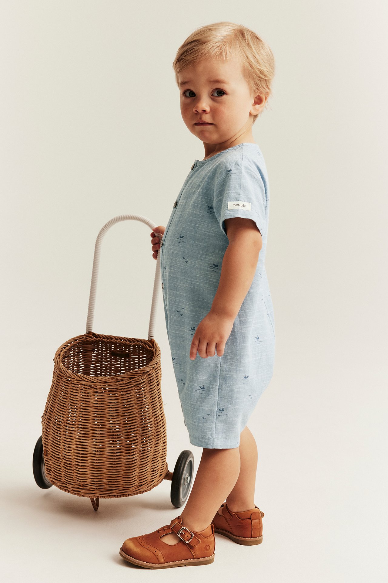 Sivukuva vauvasta: vaaleansininen valaskuvioitu puuvillahaalari, ruskeat kengät, työntäen pajukärryä.