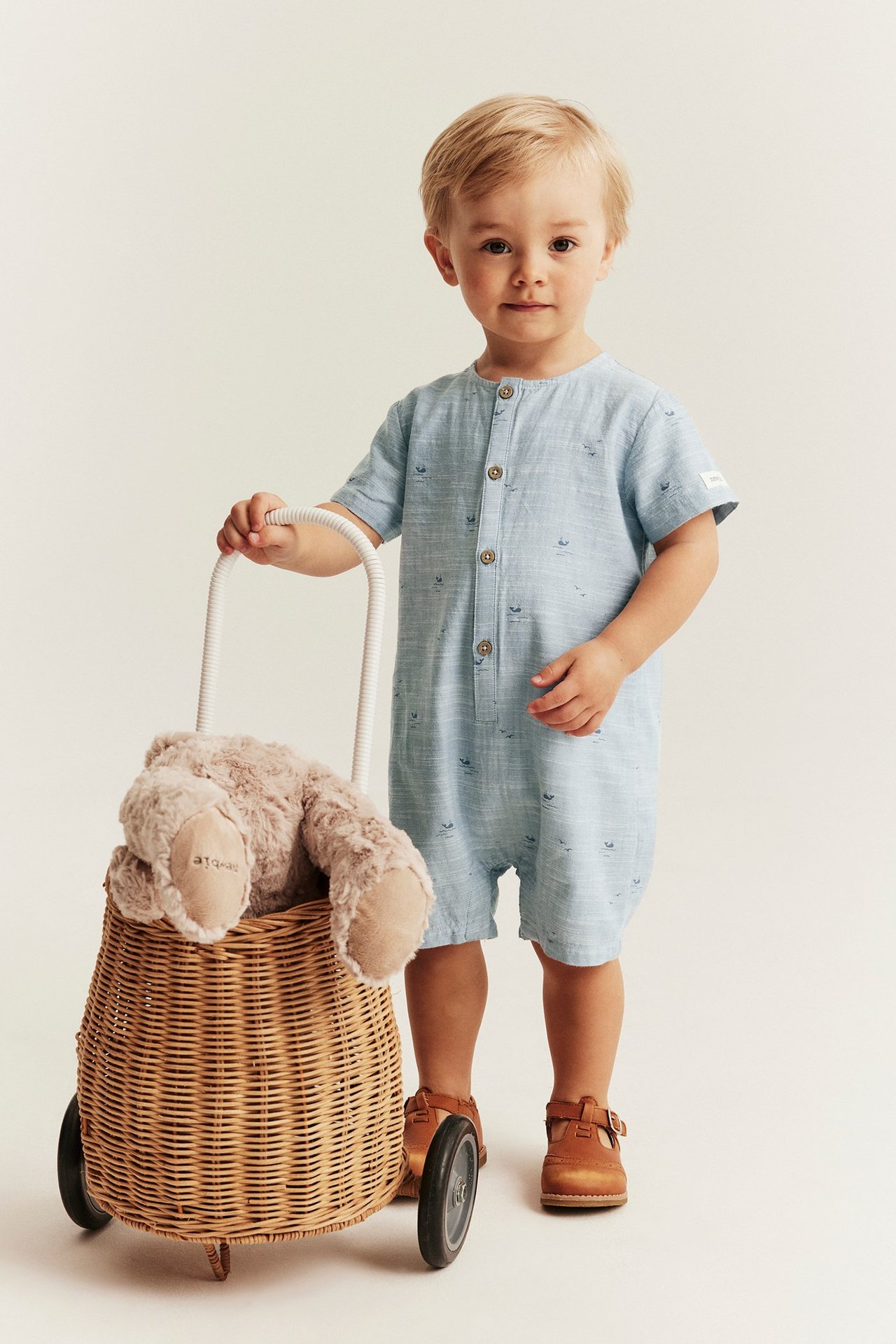 Barn i lyseblå hvalmønstret romper med knapper foran og brune sko, forfra.