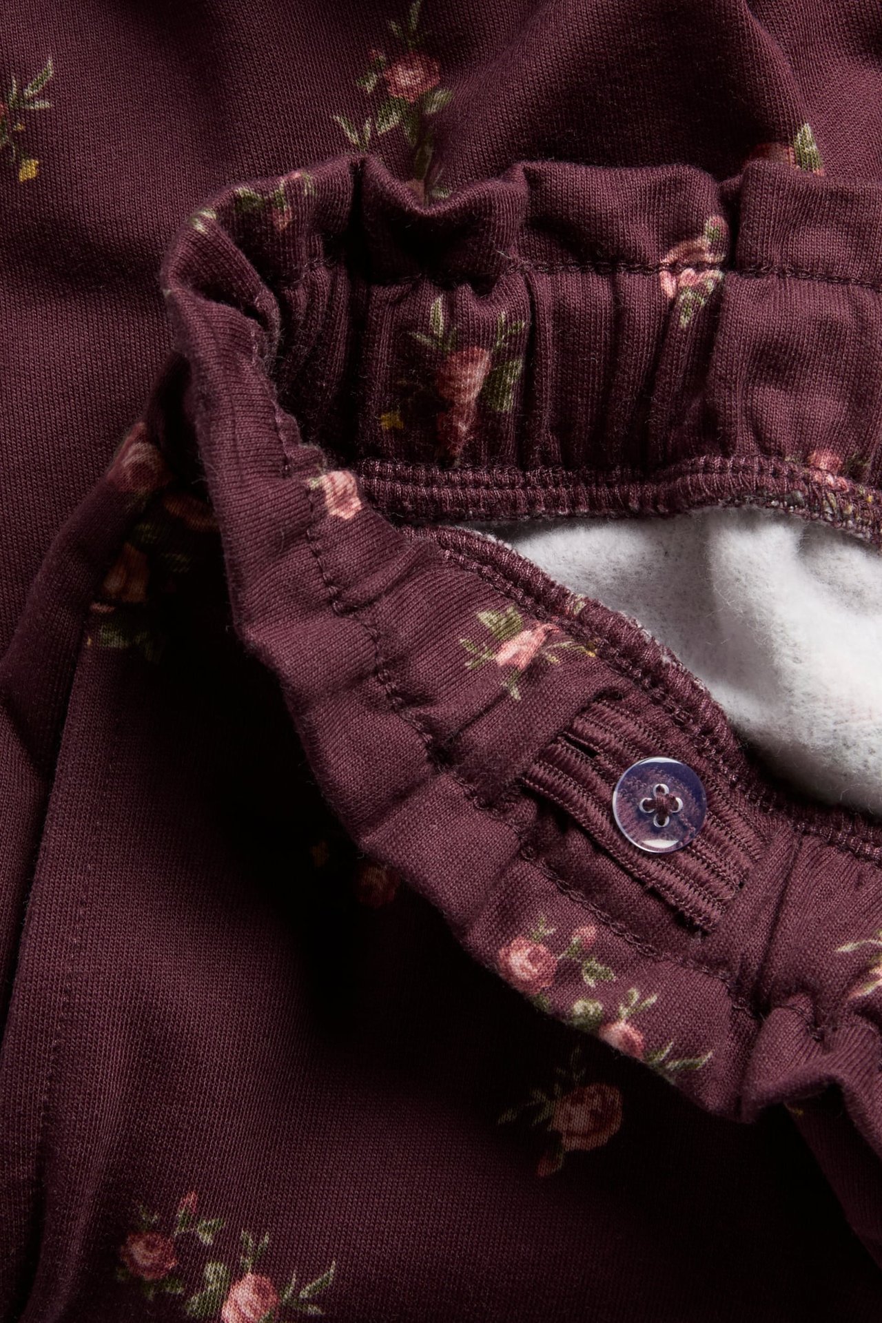 Lähikuva lasten tummanpunaisista kukkahousuista, säädettävä vyötäröresori ja violetti nappi.