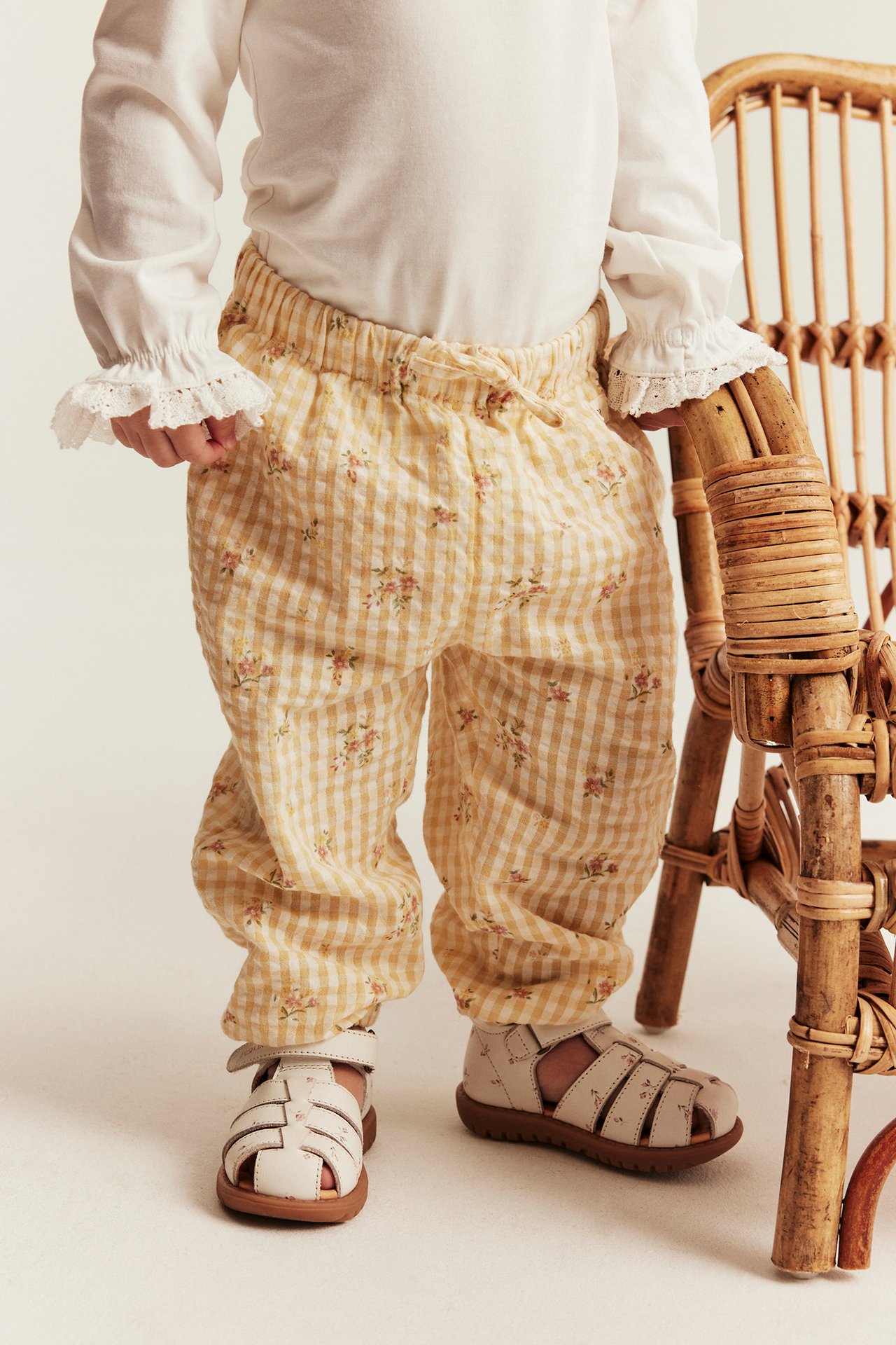 Framifrån: Ett barn bär gula rutiga blommiga vävda bomullsbyxor, en vit volangtopp och beiga sandaler.