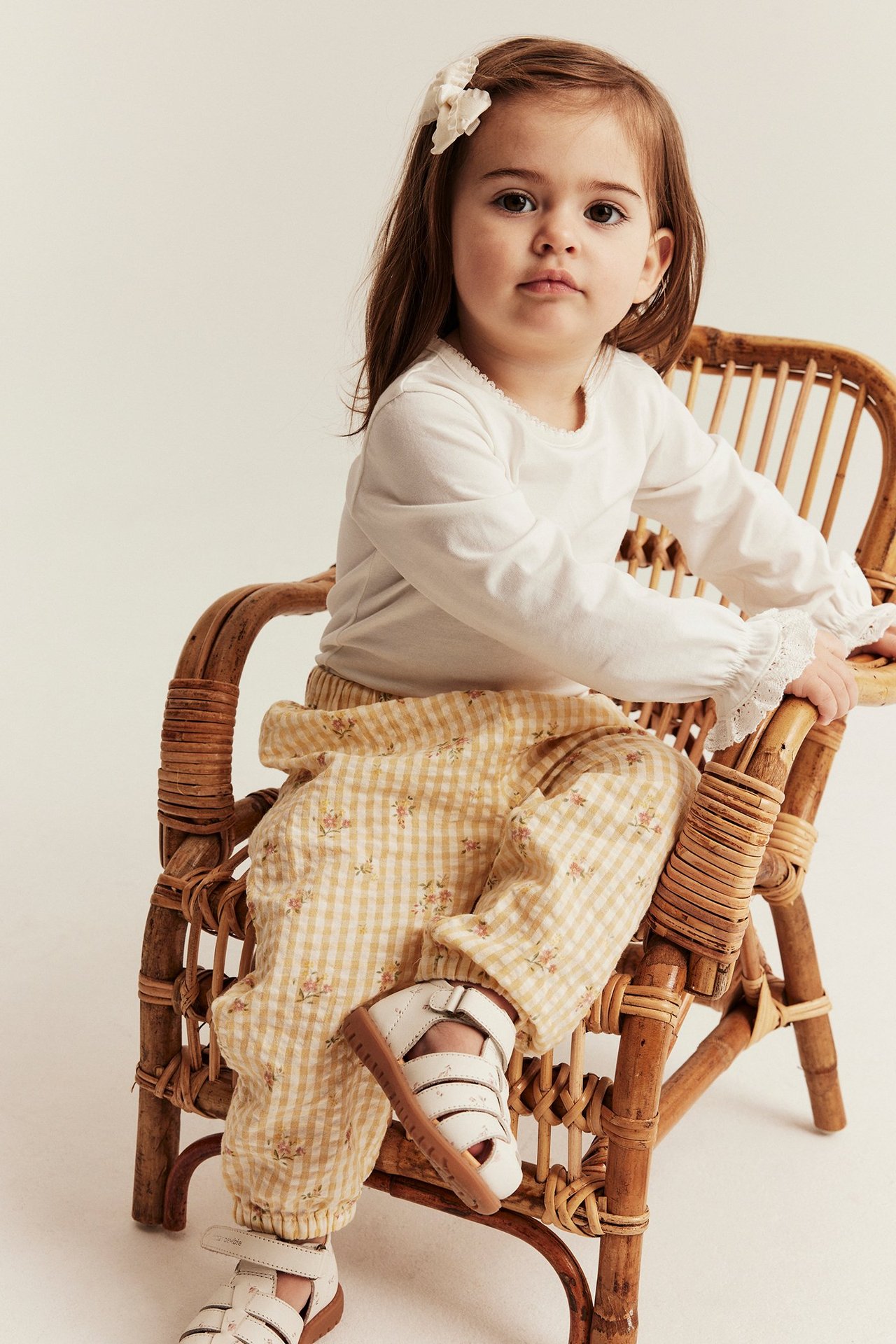 Kid i gula småblommiga rutiga vävda byxor, vit topp, vita sandaler och vitt hårspänne.