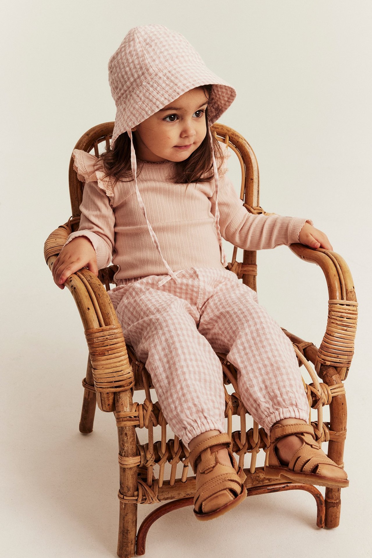 Forfra: Barn iført lys rosa rutete vevde bukser, en ribbestrikket topp, matchende solhatt og brune sandaler.