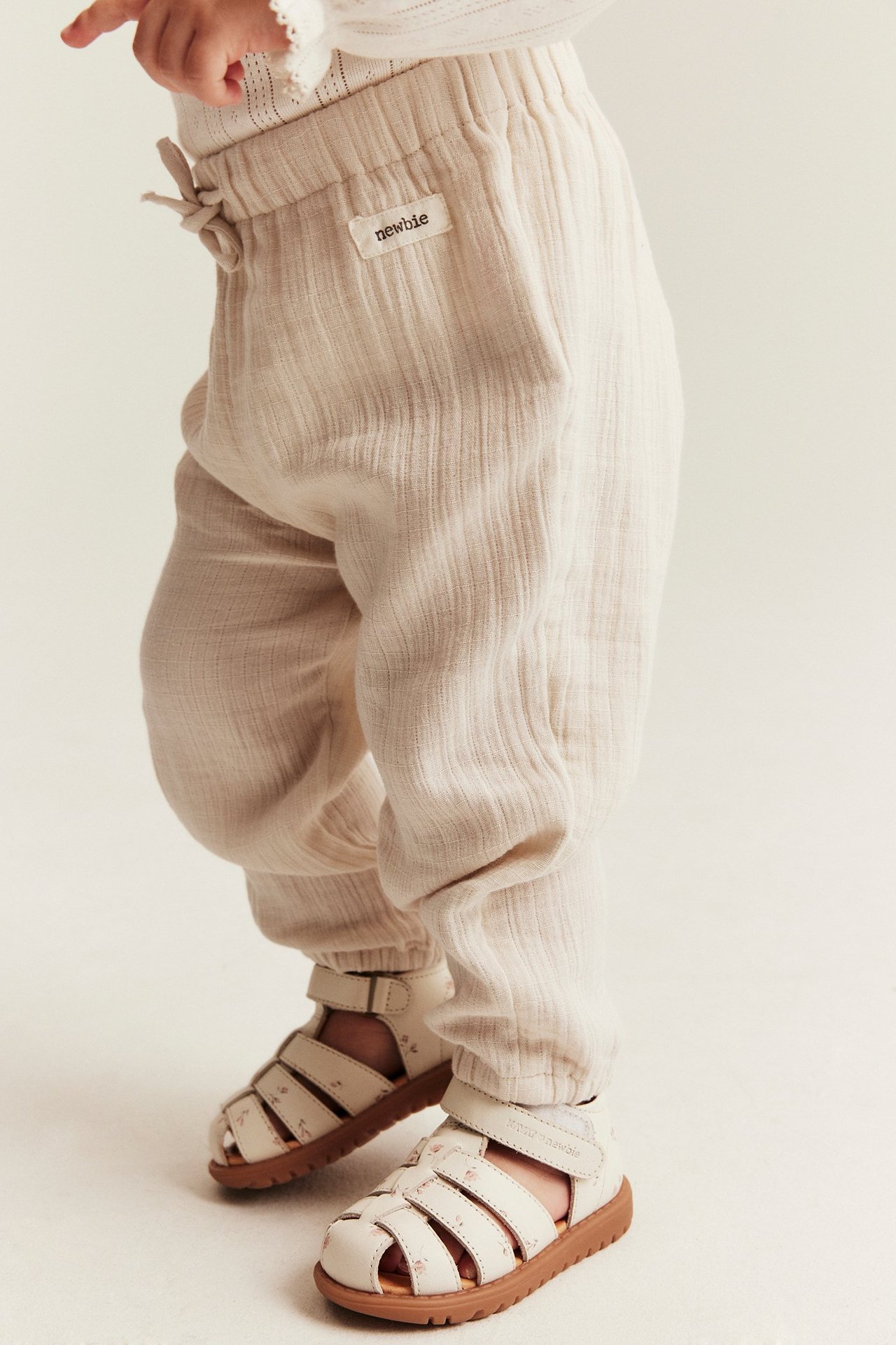 Sivukuva vauvasta vaaleanbeigessä kudotussa puuvillahousussa joustovyötäröllä ja valkoisissa kuvioiduissa sandaaleissa.