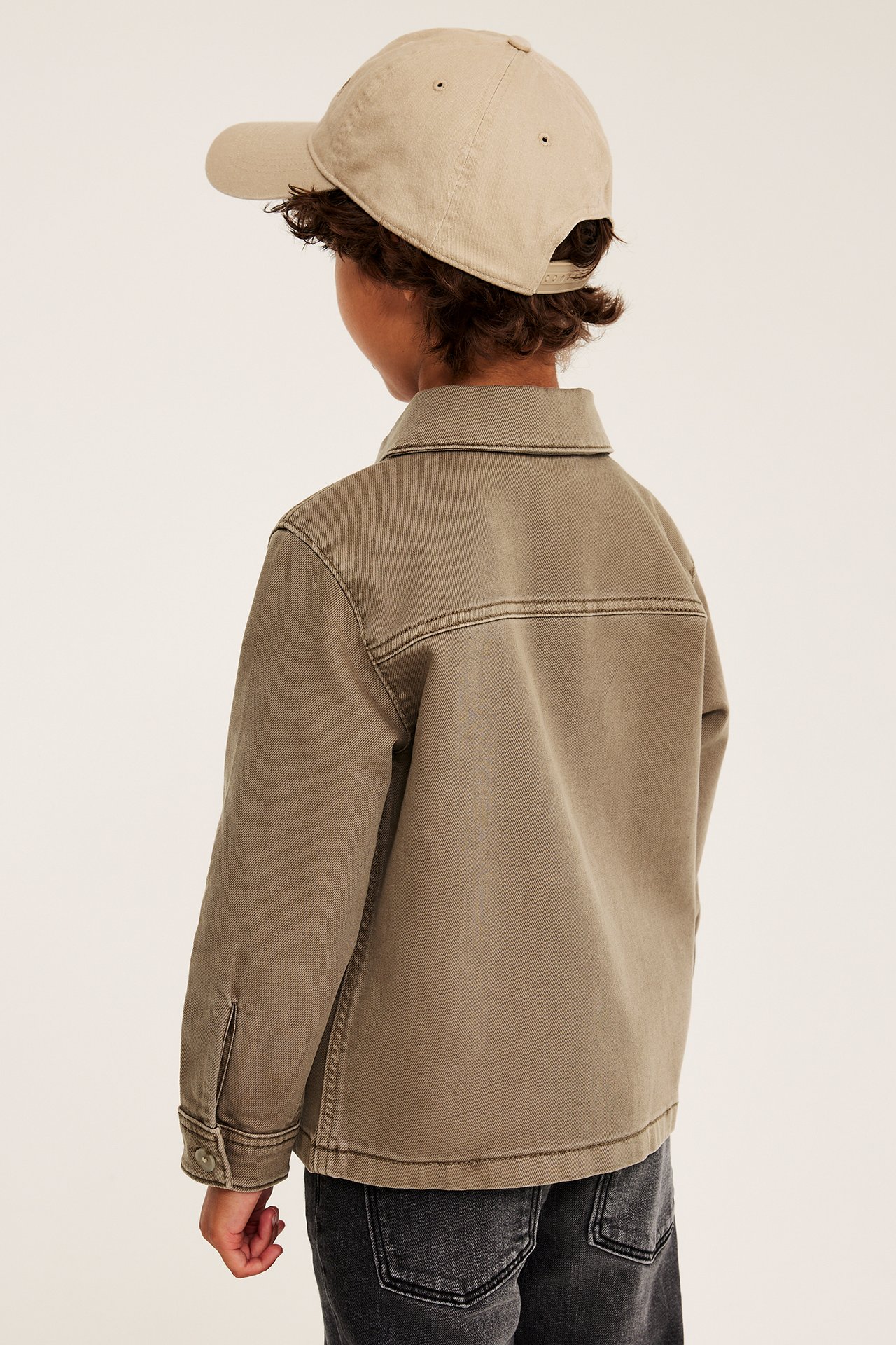 Ryggvy av ett barn i en khakigrön jeansskjorta, en beige baseballkeps och svarta jeans.