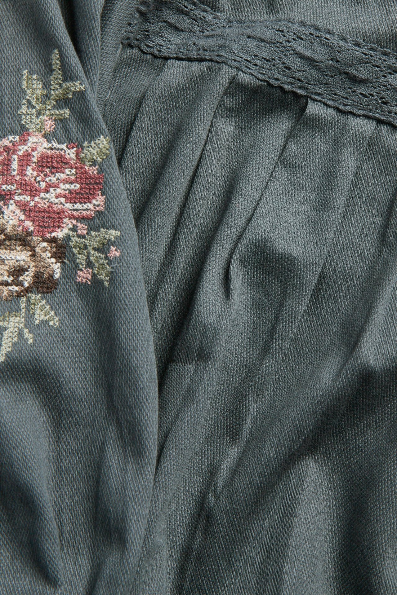 Nærbilde av et plissert mørkegrønt stoff med brodert blomstermønster og blondekant.