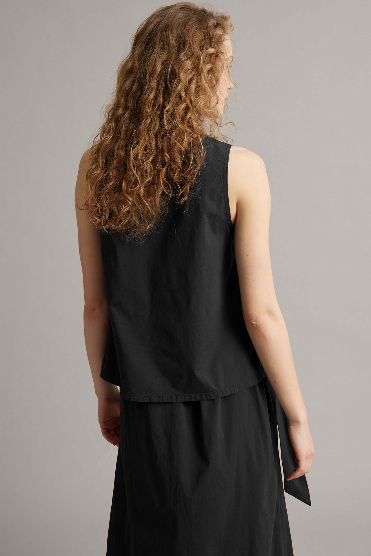 Bakifrån, kvinna i svart ärmlös linne med knytdetalj i sidan över en svart kjol.